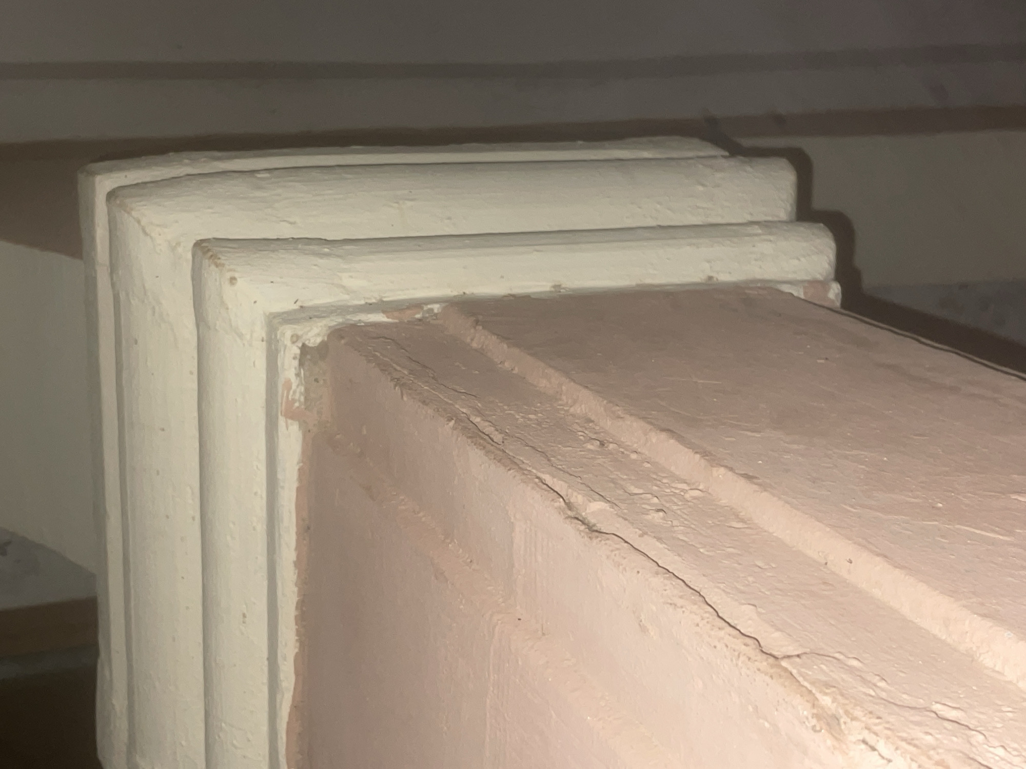 The image shows a close-up of an architectural detail, likely a ceiling or cornice. It features layers of decorative molding painted in cream and pink. The molding has a textured, possibly stucco or plaster, finish with visible brushstrokes and some minor imperfections. The cream-colored molding is layered and has a fluted or ribbed design. The pink surface beneath appears to be a structural element or a different decorative layer. The lighting suggests an indoor setting, possibly dim, with the light source casting shadows. The background is a plain, shadowed wall. The specific location in Howayi, Nigeria, cannot be determined from the visual information in the image. There are no people, animals, or specific actions depicted.