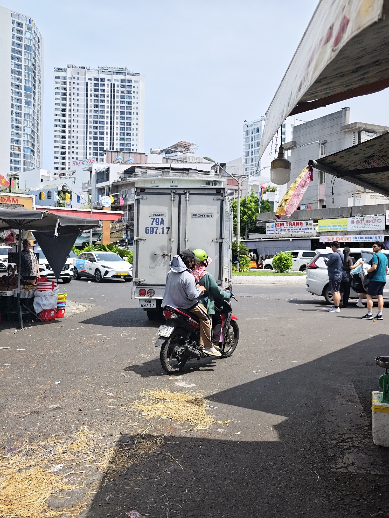 The image captures a street scene in Nha Trang Ward, Vietnam, likely a market area. In the foreground, a motorcycle carries two individuals. The driver, wearing a gray hooded jacket and a green helmet, is in focus, while the passenger behind them wears a patterned scarf and a lighter hooded garment. Scattered on the asphalt road are dry, straw-like materials, with shadows indicating direct sunlight.
Behind the motorcycle, a large white refrigerated truck with "THACO" and "FRONTIER" branding is parked. License plates are visible: "79A 697.17" on the truck and "79-M1 753.02" on the motorcycle. To the left, a market stall is set up under a black awning, displaying fruits such as bananas and what appear to be dragon fruits. A woman in a patterned top stands near the stall, and several colorful plastic buckets are stacked.
In the mid-ground, a white car with signage and a yellow license plate is stopped in traffic. Several other vehicles, including a white sedan and a darker car, are also visible. The background features modern high-rise buildings, suggesting an urban environment adjacent to the market. The sky is clear and bright. The overall impression is a bustling daytime street scene in a Vietnamese city.