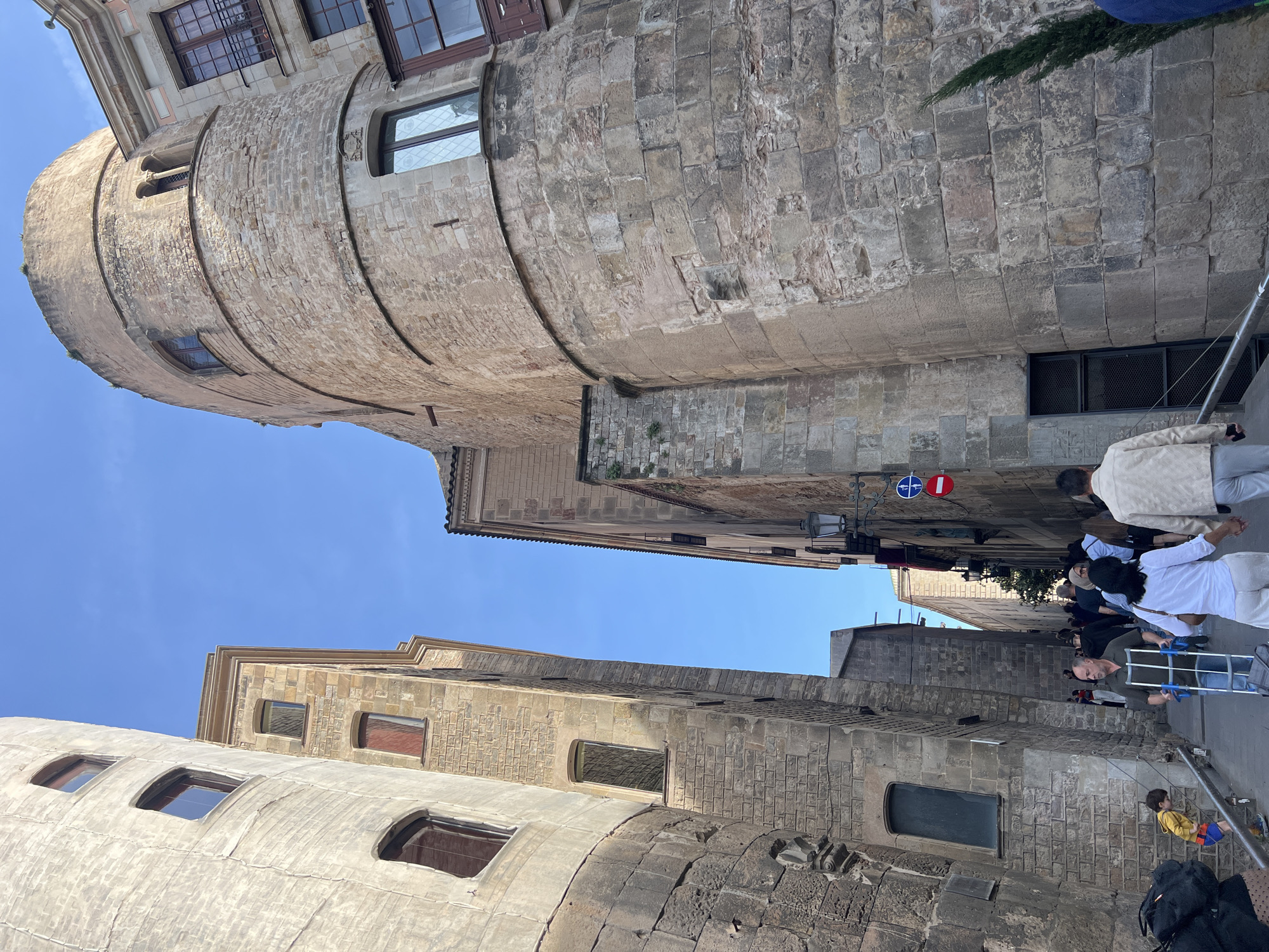 The image captures a sunlit, narrow street scene in a historic European city, specifically Barcelona, characterized by ancient stone architecture. The perspective is looking upwards between tall buildings, revealing a clear blue sky.
On the left, a large, rounded stone structure, resembling an ancient tower or bastion, dominates the mid-ground, featuring several rectangular and arched windows set into its weathered surface. Another similar but less distinct rounded stone structure is visible in the left foreground. Opposite this, on the right, is another tall building with a flat stone facade and square windows, some with decorative grates. Parts of this building also show signs of ancient stone block construction.
Several people are visible on the street below, engaged in everyday activities. A man in a dark shirt is pushing a metal shopping trolley or cart. Other pedestrians are walking along the street. A small child in a yellow shirt and blue shorts sits by the side of the road on a low structure.
Notable details include two circular street signs attached to a pole on the right building: one is a blue circle with a red diagonal line and red border (indicating no stopping or no parking), and the other is a red circle with a white horizontal bar (indicating no entry or do not enter). A street lamp is also visible on the right building. The daylight is bright, suggesting a clear day, likely late morning or afternoon, with some shadows cast by the buildings. The overall impression is a bustling yet historic street, blending ancient Roman or medieval structures with modern urban life.