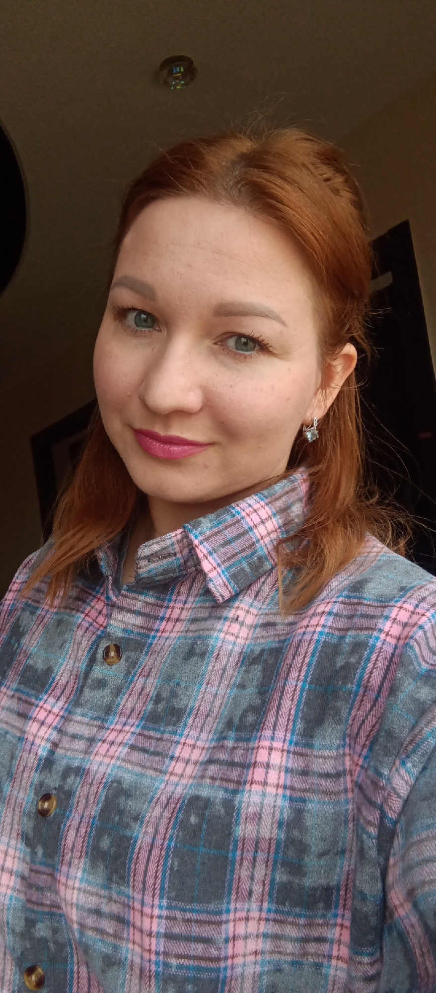 A medium close-up, selfie-style portrait features a woman with reddish-brown hair and green eyes, looking directly at the camera. She has light skin, some faint freckles, and is wearing pink lipstick. Her eyebrows are well-defined, and her eyelashes appear long. She wears a plaid shirt with a pattern of pink, greyish-blue, and light blue lines. A small, square-shaped silver earring with a light-colored stone is visible on her right ear. Her hair is parted and appears to be pulled back or to the sides, with some loose strands framing her face.
The background is mostly dark and out of focus, suggesting an indoor setting. A dark doorway or frame is visible on both sides of the woman. Above her head, a small, circular, ceiling-mounted object, possibly a light fixture or smoke detector, is visible, showing some colorful reflections. The woman has a slight smile, conveying a pleasant expression. There is no visible text in the image.