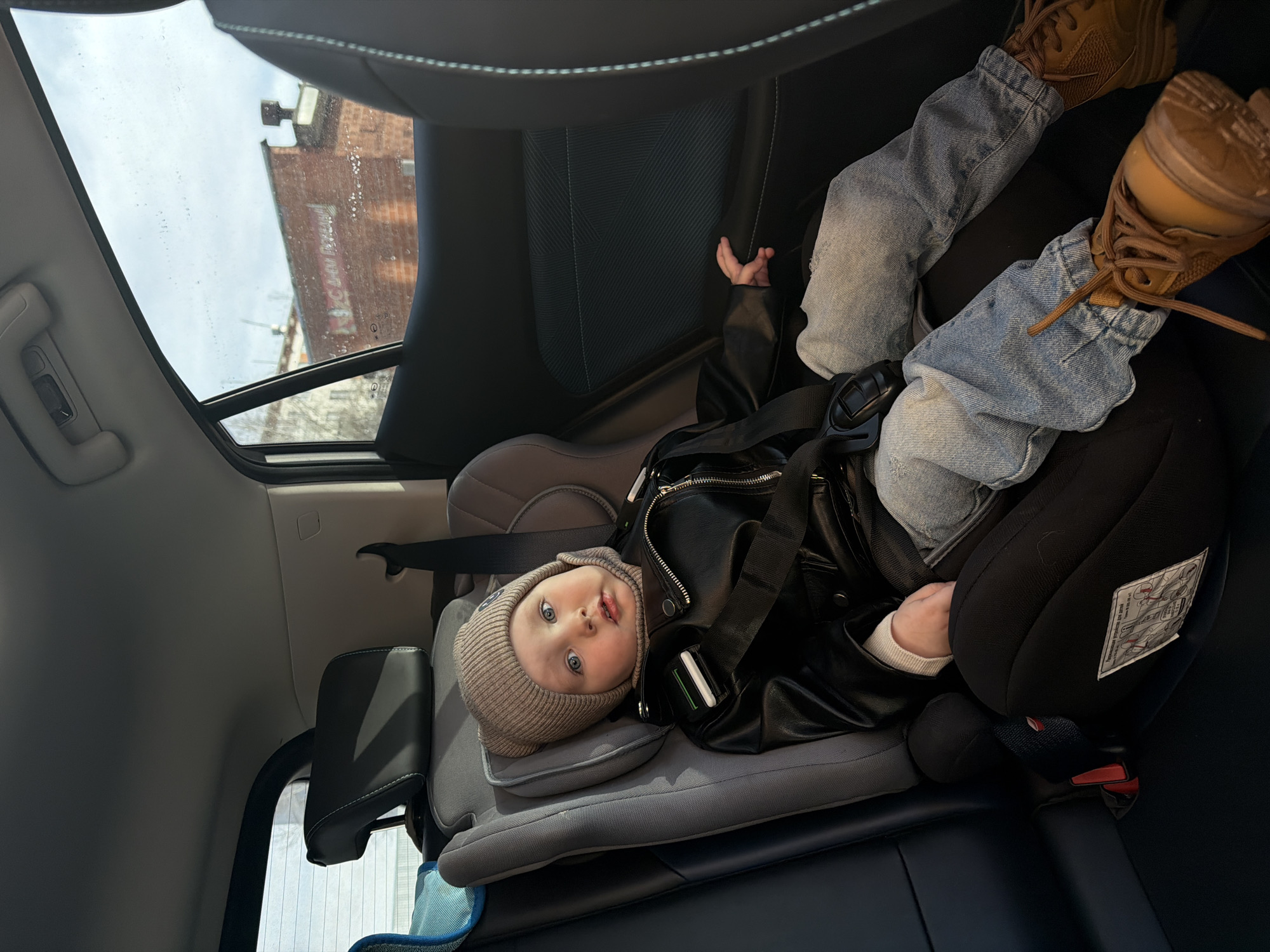 A young child, secured in a car seat within a vehicle, appears to be gazing directly at the camera. The child is dressed in a brown knit hat, a black leather jacket, blue jeans, and brown boots. The vehicle is positioned in Chaikovsky, Russia, as indicated by the reflection of a building outside the window. The building's facade suggests a typical urban structure found in the city. The child is facing the camera, and a hand is visible resting on the side of the car seat. The car seat is properly installed in the car. The rest of the car's interior is dark.