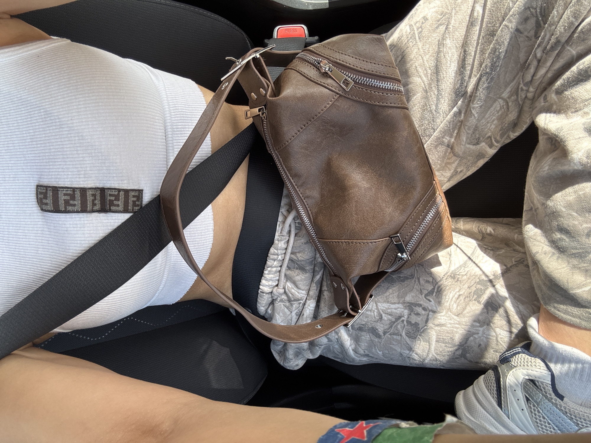 The image shows a person sitting in the driver's seat of a vehicle, wearing a white ribbed tank top with a dark brown logo patch on the chest, secured by a black seatbelt. A brown leather bag is draped across their lap. The person is wearing camouflage pants and a white and grey sneaker is visible. There is a partial view of a tattoo with a red star and green background on their leg. The seatbelt buckle is visible in the background. The location appears to be inside a car, but specific details about the city of Riverside, United States of America are not identifiable from the image.