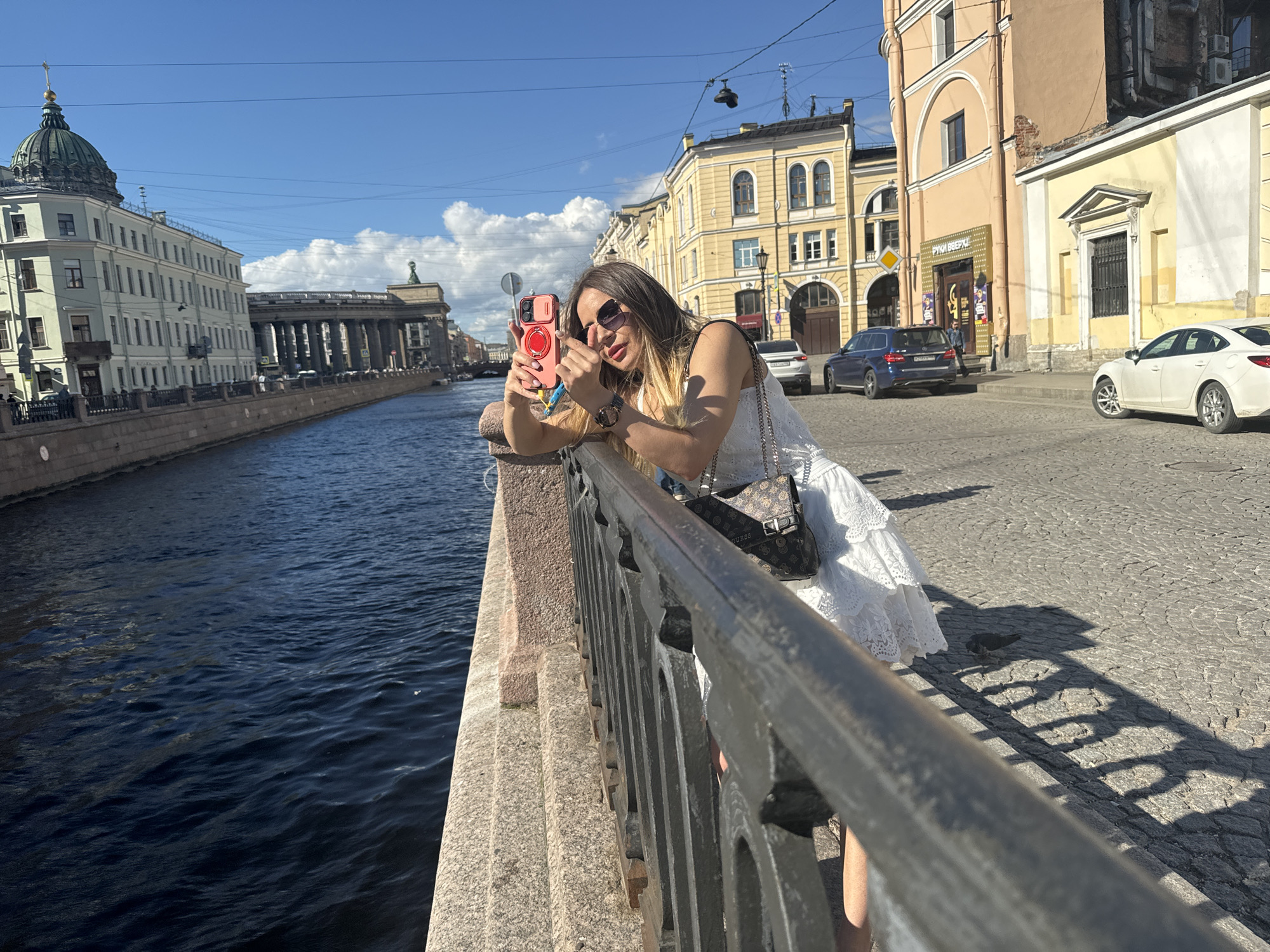 A woman in a white dress and sunglasses is taking a selfie with her pink smartphone. She is leaning over a metal railing beside a canal. The canal's dark water reflects the blue sky and white clouds. In the background, there are several buildings, including a large building with a green dome and a long colonnade. The sun is shining brightly, casting shadows on the cobblestone street behind the woman. The overall mood is relaxed and summery. The scene appears to be in Saint Petersburg, Russia, as suggested by the architecture. The time of day is daytime, likely midday, given the bright sunlight and clear sky.