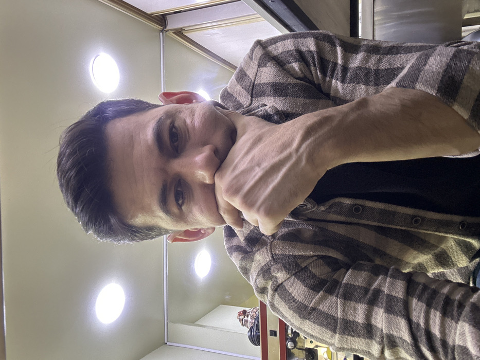 A man in a brown and beige plaid flannel shirt poses indoors. His left hand is raised to his mouth in a pensive gesture. He looks directly at the camera with a serious expression. The lighting in the room is bright, with several visible ceiling lights. The background includes parts of a machine or device, and the interior walls are a light beige color.