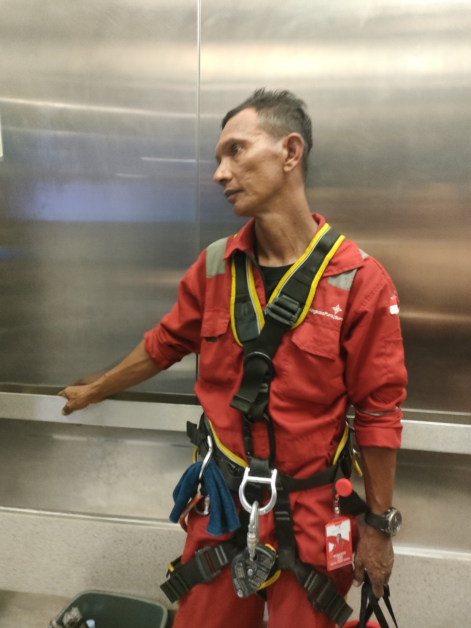 A man wearing a red jumpsuit and a safety harness is standing in an elevator. The elevator's walls are made of stainless steel. The man has short, dark hair and is looking to his right. He is holding onto the elevator door with his left hand. A blue cloth and a lanyard with an ID badge are attached to his harness. He is also wearing a watch on his right wrist. The image appears to have been taken in an indoor setting, likely within a building in Pekanbaru, Indonesia, given the context provided.
