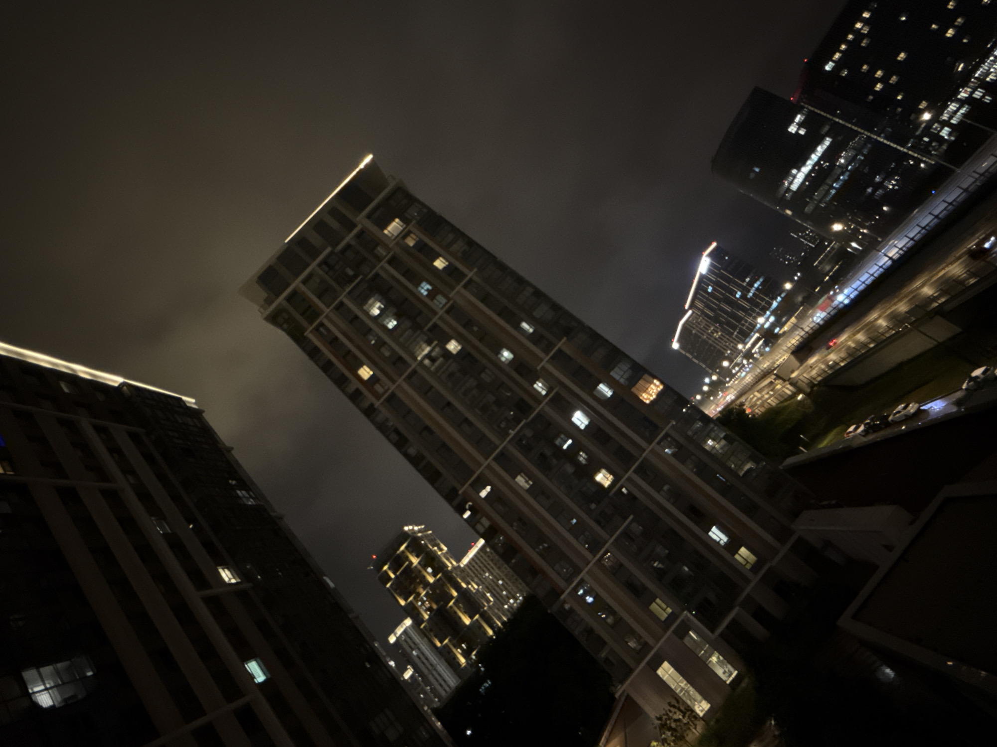 The image depicts a nighttime aerial view of a modern city district in Khimki, Russia, characterized by towering, illuminated skyscrapers and a street with vehicle traffic. The perspective is angled, giving a dynamic feel to the urban landscape.
Dominating the foreground and mid-ground are several large, rectangular apartment buildings and office blocks. Their facades are adorned with numerous windows, many of which are lit, suggesting occupancy or internal lighting. Some of these buildings have distinctive architectural features, such as balconies or external structural elements. One prominent building in the center features a strip of bright, white light running along its uppermost edge, highlighting its height and modern design. Another building to the left, partially obscured by a darker structure, also shows a luminous edge.
In the distance, more buildings are visible, their lights creating a bokeh effect against the dark, cloudy sky. A street or highway runs through the scene, with streaks of light from moving vehicles indicating traffic flow. The wet surface of the road reflects the city lights, suggesting recent rain or damp conditions.
The overall impression is one of a bustling, contemporary urban environment at night, with a focus on architectural lighting and the expansive nature of the city. There are no visible individuals or specific interactions. The environment is predominantly man-made, consisting of architecture and infrastructure.