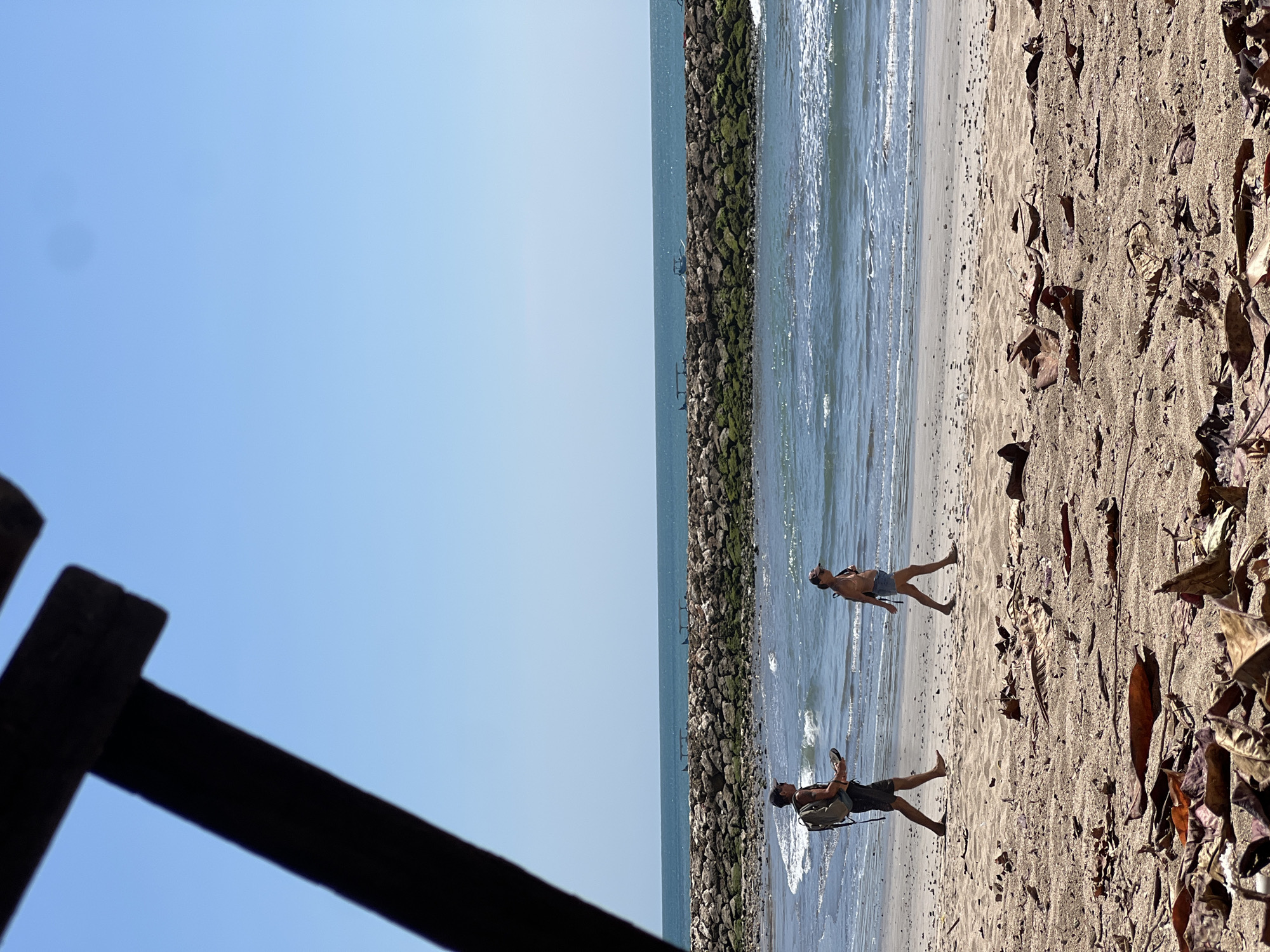 The image depicts a beach scene in Kuta, Indonesia, under a clear blue sky. In the foreground, dried leaves are scattered on the sand. Two individuals are walking along the shoreline. A man, wearing a backpack, walks to the left, holding flip-flops. A woman, in a bikini top and denim shorts, walks to the right. The ocean is visible behind them, with waves gently breaking on the shore. A long breakwater, constructed of rocks and covered in green moss, extends into the sea. Several small boats are visible near the breakwater. The overall scene is tranquil, capturing a typical day at the beach.