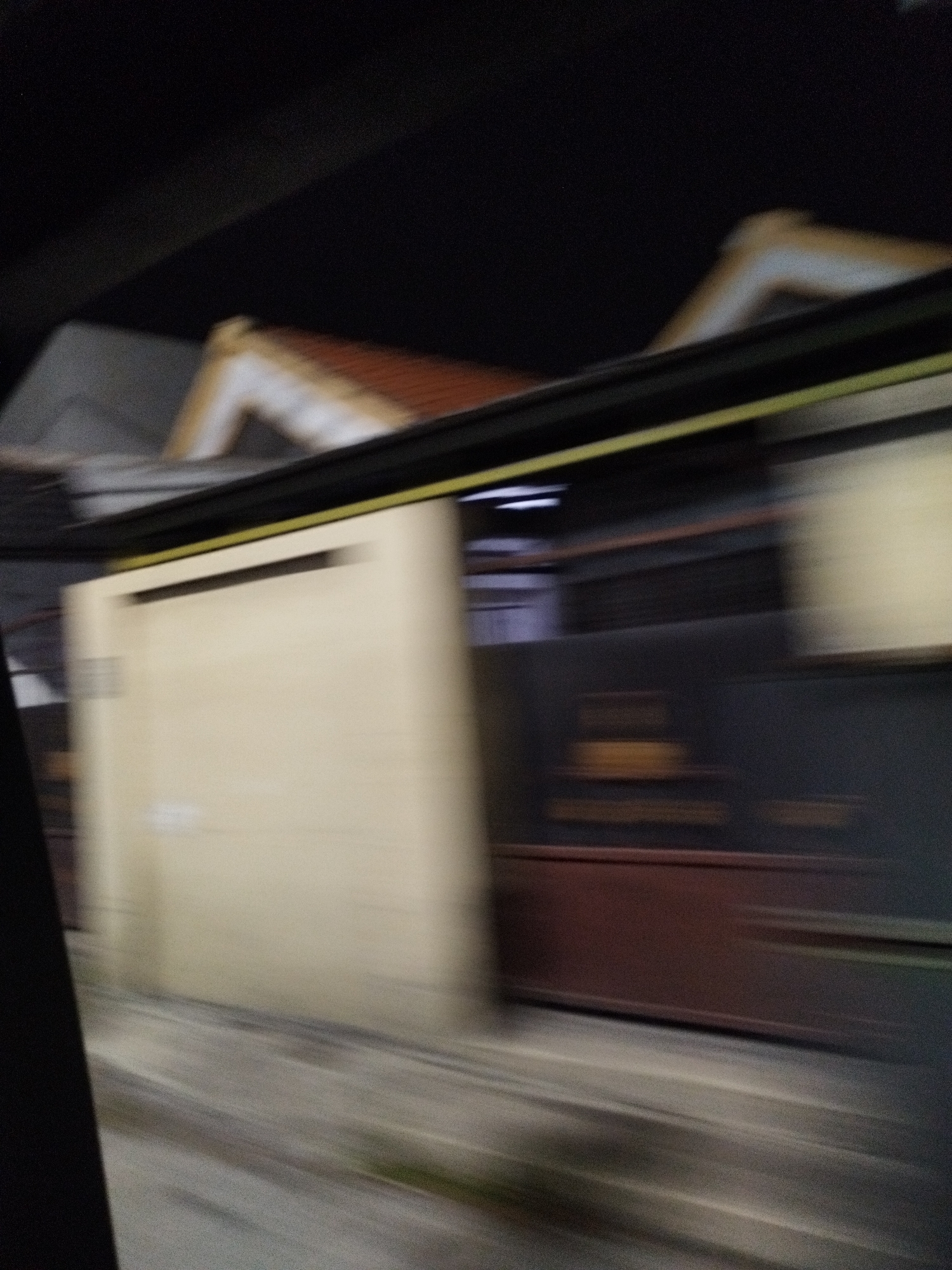 The image captures a blurred nighttime scene of a building in Surabaya, Indonesia, likely viewed from a moving vehicle. The structure features a light-colored wall on the left side, possibly a garage door or a large panel, with a dark rectangular opening near the top. To the right, a darker wall or structure is visible, with horizontal bands of lighter and darker colors. Above these walls, a sloped roof with reddish-brown tiles is discernable, partially obscured by a dark, possibly asphalt, overhead structure. The overall impression is one of speed and motion, with light streaks suggesting streetlights or reflections. The low-light conditions and motion blur make precise identification of architectural details challenging, but the scene suggests an urban environment at night.