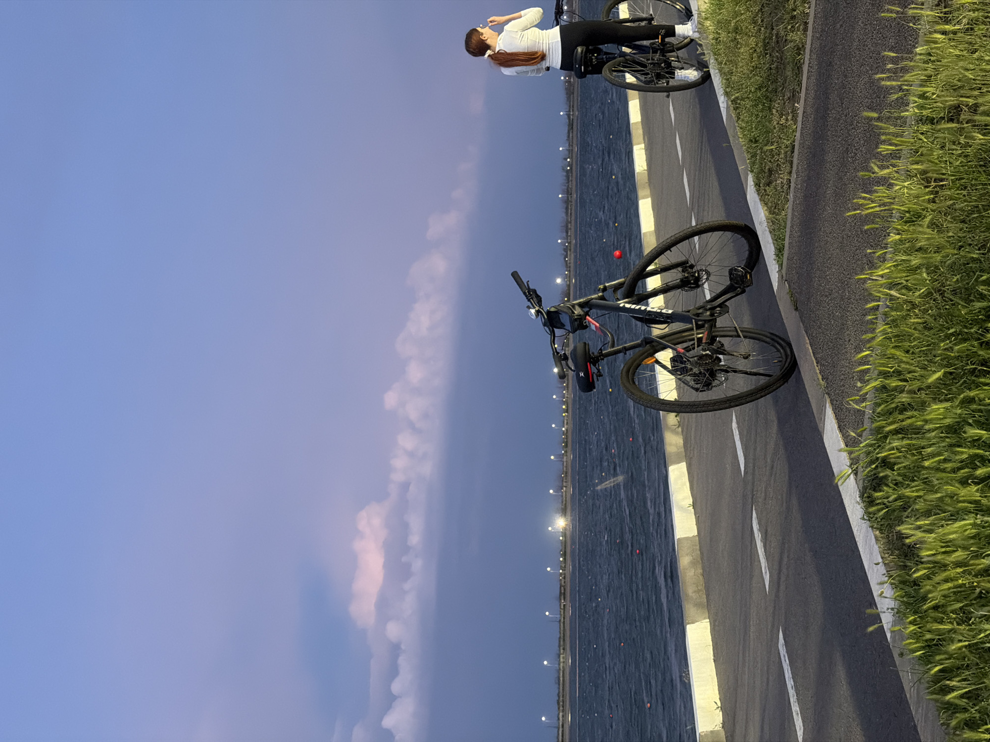 A bike path in Rostov-on-Don, Russia, is captured in this image. A woman in a white top and dark pants stands near two bicycles. The path borders a body of water with lights and markers visible in the distance. Grass grows along the edge of the path. The sky is a blend of blue and purple hues, suggesting either dawn or dusk.