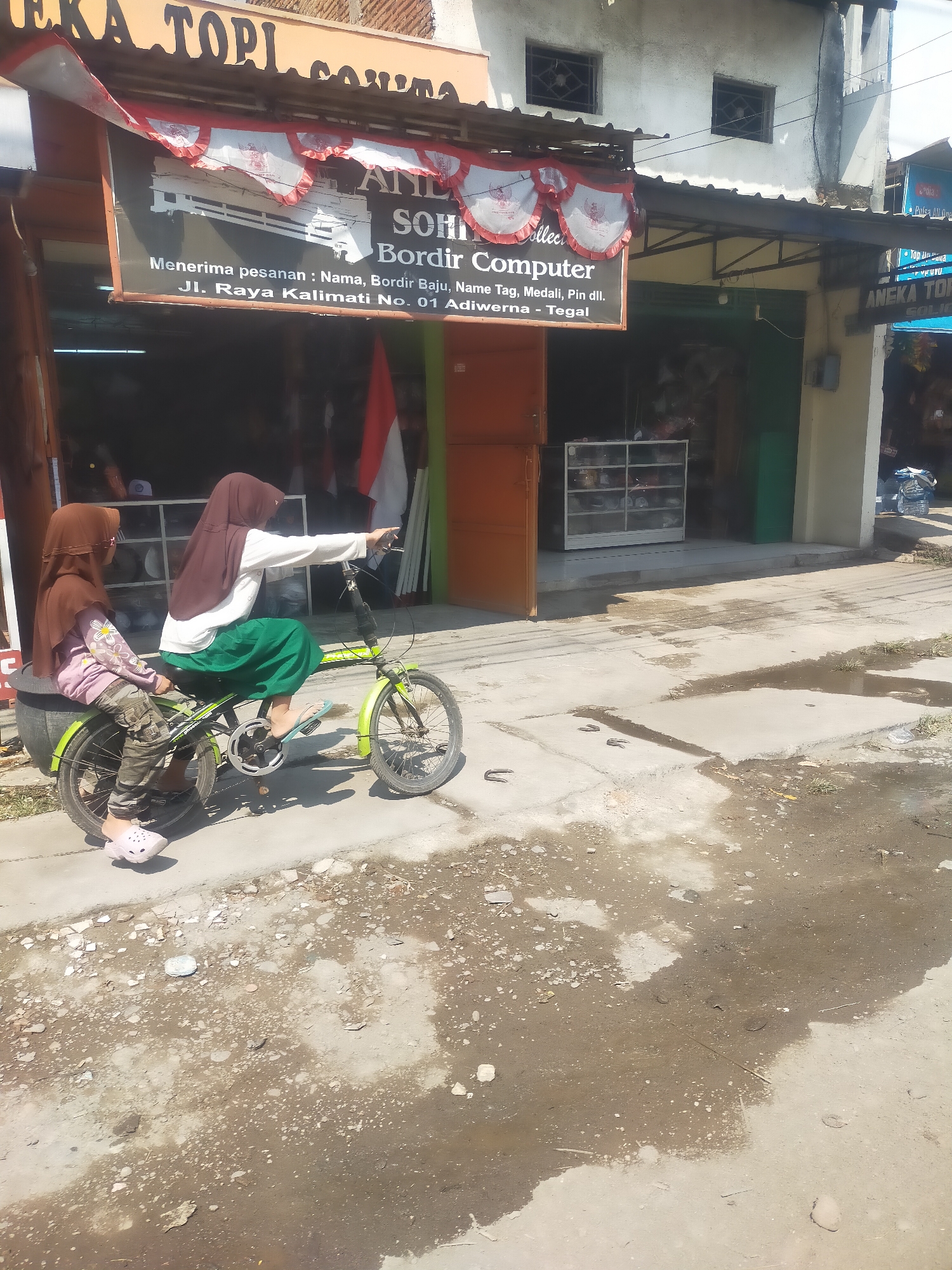 Two young girls wearing hijabs are riding a small, green bicycle on a paved street in front of a shop in Lemahduwur, Indonesia. The girl in the front is wearing a brown hijab and a pink shirt with flower embroidery, and is sitting behind the other girl. The girl in the back is wearing a dark brown hijab and a white long-sleeved shirt, and is pedaling the bicycle. She is also wearing a long green skirt. The bicycle has a black basket attached to the rear. The shop behind them has a sign that reads "SOHI Bordir Computer" and lists services for name tags, embroidered clothing, and other items. The shop also has a display cabinet inside. The street is wet in patches, suggesting recent rain. The area appears to be a commercial district with buildings lining the street.