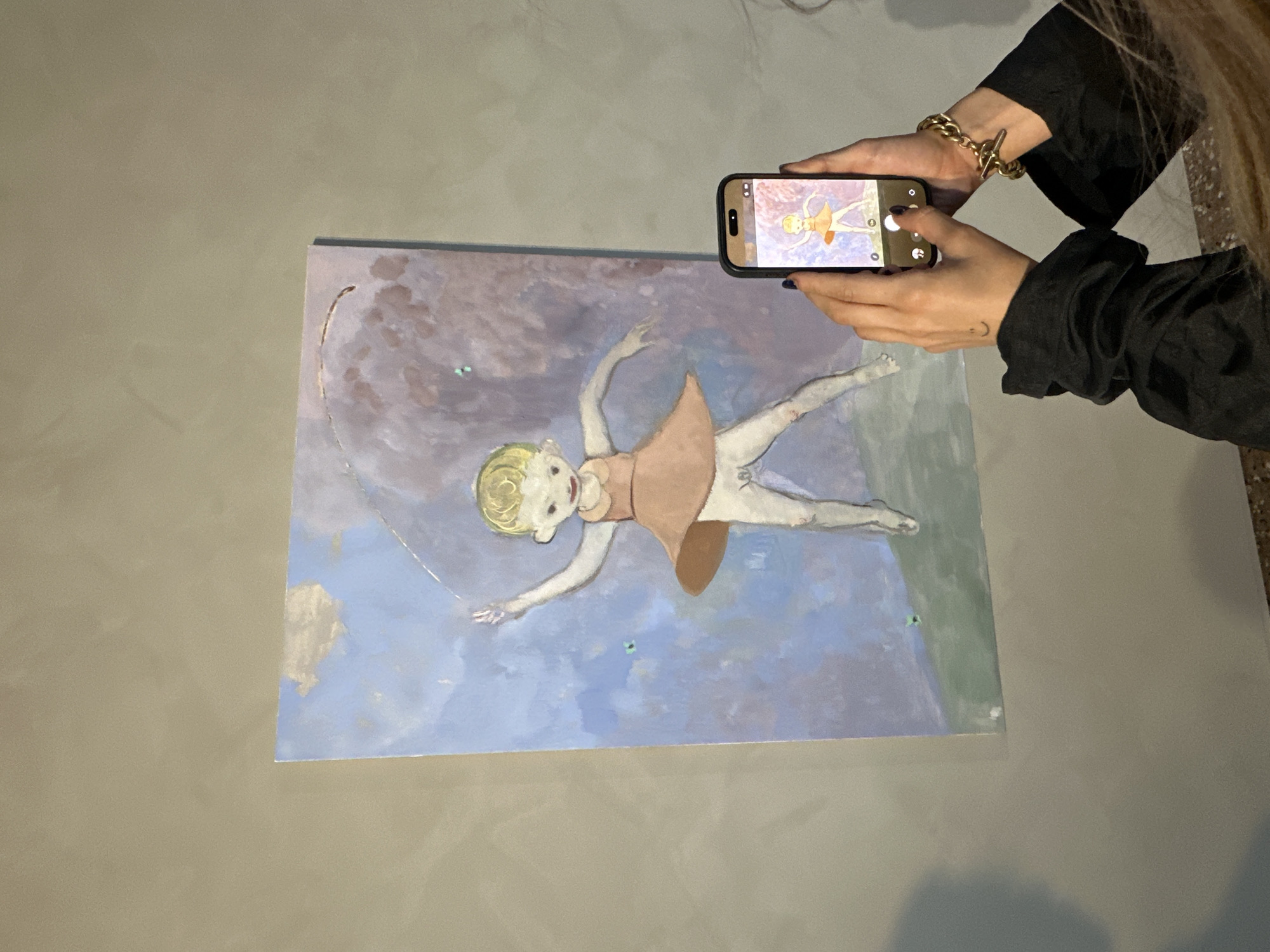 The image shows a person holding a smartphone to photograph a painting displayed on a gray wall. The painting depicts a figure resembling a child with blonde hair and a light pink dress. The figure is positioned as if floating or dancing against a backdrop of blue and purplish hues. A curved line, possibly representing a rope or ribbon, extends above the figure. The smartphone in the image is actively displaying the painting, mirroring the artwork being photographed. The person holding the phone is wearing a dark, long-sleeved shirt and a gold bracelet. The background is blurred, suggesting the setting might be a gallery or exhibition space. There is no specific information in the image to confirm it is in Venice, Italy.