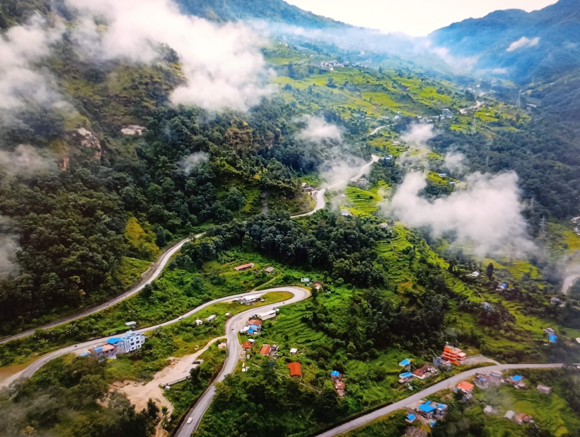 This is an aerial, high-angle, long shot of a winding mountain road cutting through a lush, green, terraced landscape. The road is paved and appears to have multiple lanes in some sections, with several small buildings and houses scattered along its path. Patches of white clouds drift through the scene, obscuring parts of the mountains in the background. The vegetation is dense and vibrant green, indicating a tropical or sub-tropical environment. There are no visible people or animals in the image. The scene conveys a sense of remote beauty and natural grandeur. There is no indication that this location is in the city of Dubăsari town council, Moldova.
