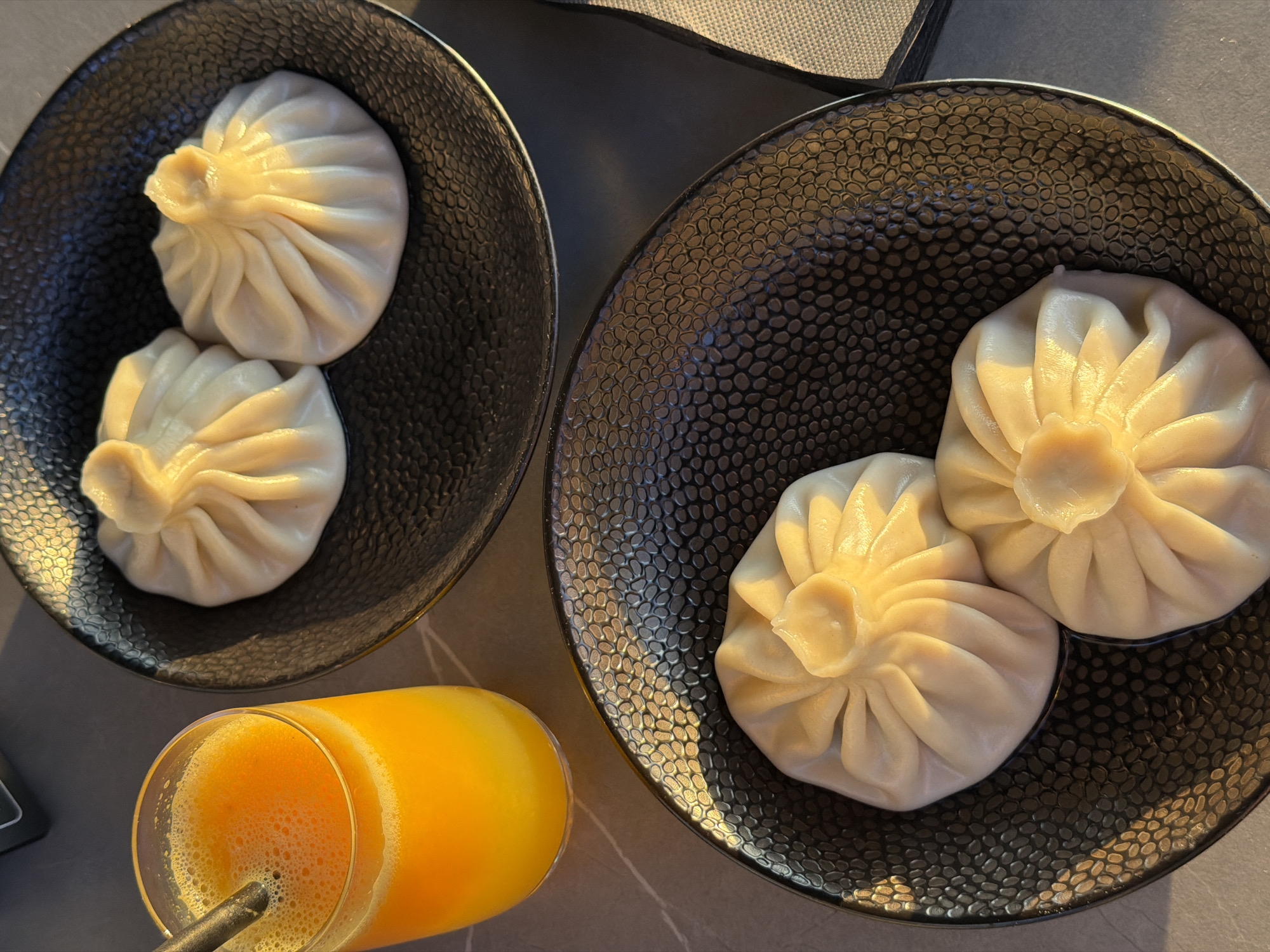 Two dark plates hold dumplings, two per plate. A glass of orange juice with a straw sits near one plate. The setting is likely a restaurant table in Rafailovići, Montenegro.