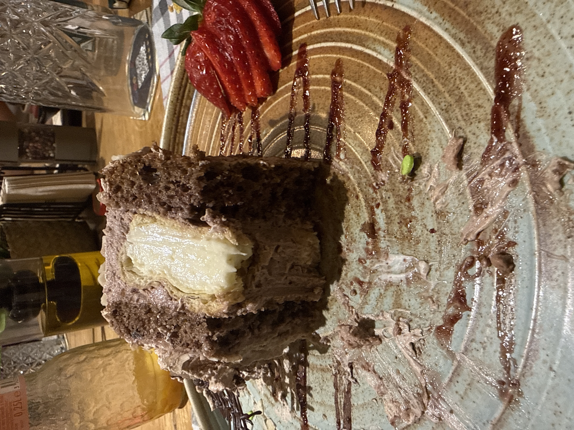 The image depicts a partly-eaten chocolate cake with a creamy filling on a patterned plate. The cake is served with sliced strawberries. Dark syrup is drizzled on the plate. A fork is visible next to the cake. In the blurred background, there are bottles, glassware, and possibly spices. The image is likely taken indoors, possibly in a restaurant, but the location within Sofia, Bulgaria cannot be determined from the visual data.