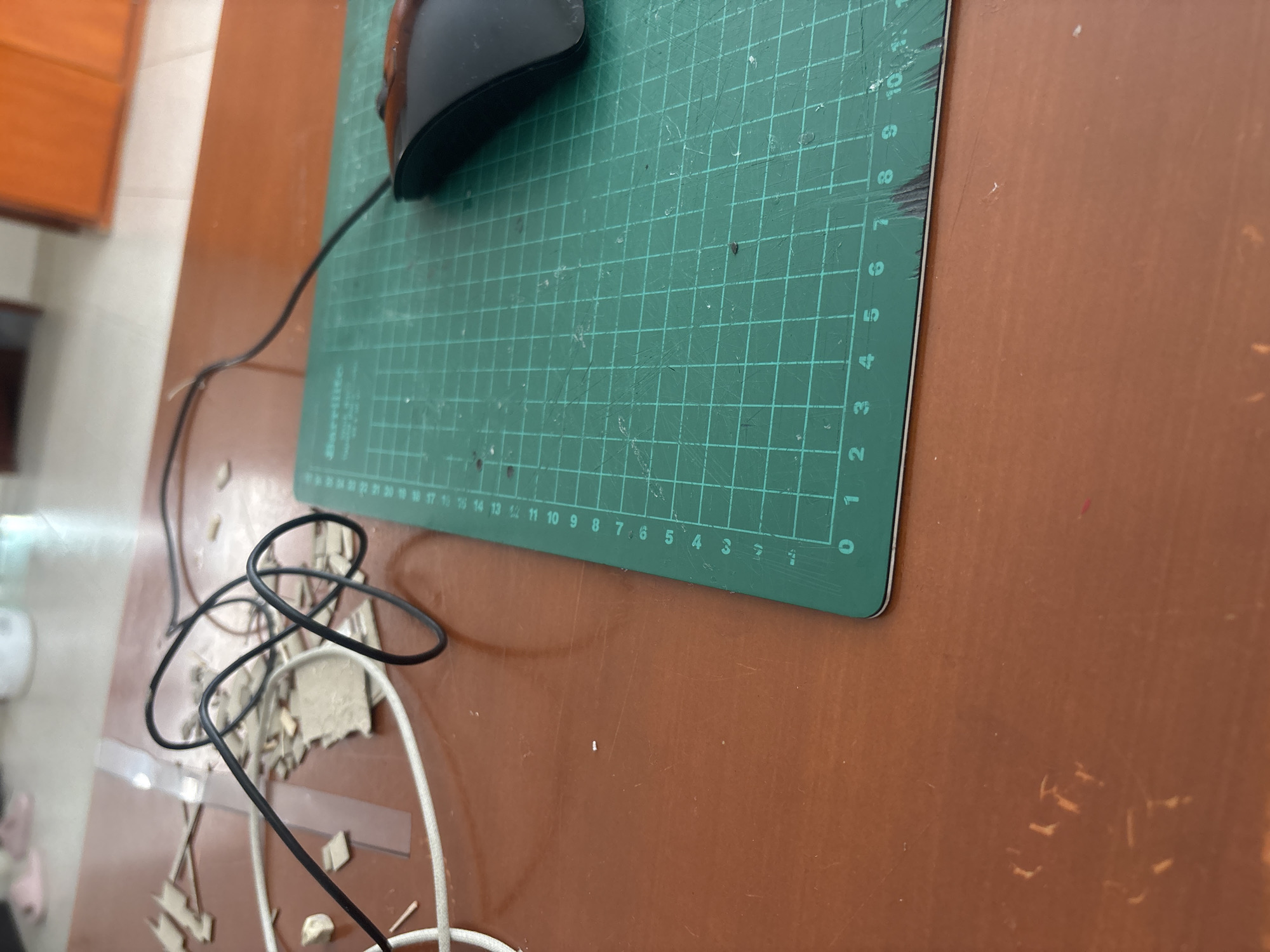 The image depicts a desk setting. A black computer mouse rests on a green cutting mat, which is marked with grid lines and numerical measurements. A black cord connects to the mouse and is coiled on the desk alongside a white cord. Debris from crafting materials, possibly cardboard or paper, is scattered around the cords. The desk has a smooth, wood-like surface and a reddish-brown color. In the background, a portion of a wooden cabinet is visible. The image provides no indication of the location of San Pablo del Monte, Mexico.
