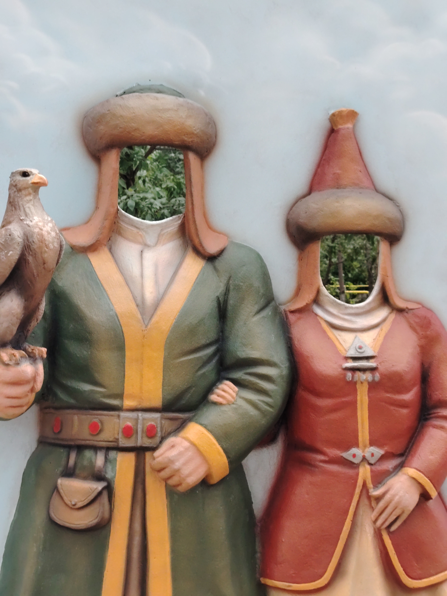 The image shows a cut-out prop designed for tourists, depicting two figures in traditional attire, a male figure on the left and a female figure on the right. The prop is made of sculpted material, likely fiberglass or plaster, painted in vibrant colors. The male figure is dressed in a green tunic with yellow trim, a wide belt with red accents, and a fur-lined hat. He is holding a sculpted falcon on his left arm, and his right arm is around the female figure's waist. The female figure wears a red tunic with yellow trim and a conical hat. Both figures have cutouts for faces, allowing visitors to pose for photographs. The prop is set against a light blue background with white cloud-like patterns. In the background, visible through the face cutouts, is a blurry green landscape with trees, suggesting an outdoor setting. This type of interactive photo prop is commonly found in tourist areas. The style of the clothing suggests a historical or cultural theme, possibly related to nomadic peoples of Central Asia. Given the prompt's specific mention of Astana, Kazakhstan, this prop is likely located in a park, cultural center, or tourist attraction within the city, intended to offer visitors a playful way to engage with local heritage.