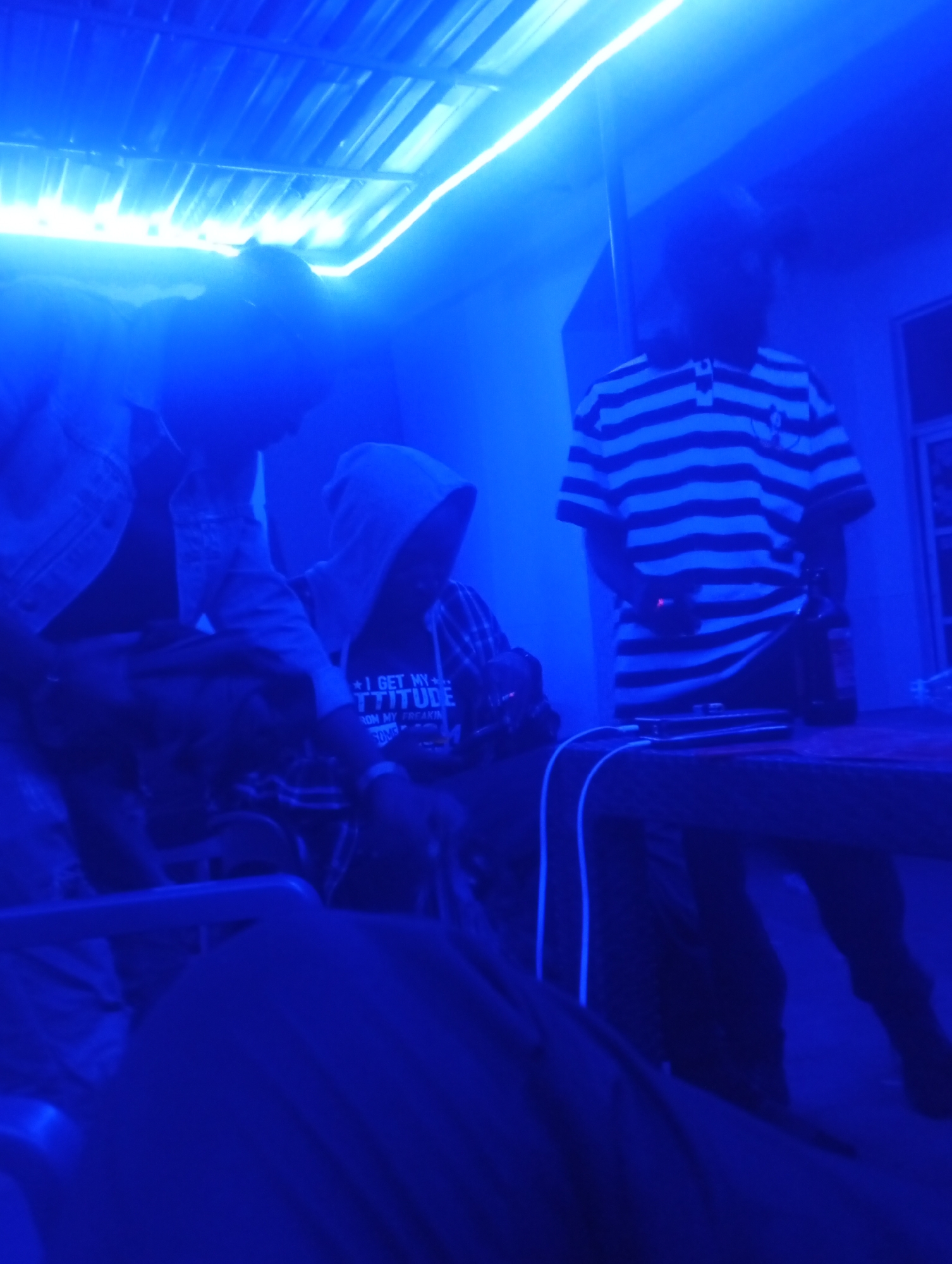 This image depicts an indoor night scene bathed in intense, artificial blue light, likely from LED strips along the ceiling, suggesting a bar, lounge, or party environment in Barakin Akawo, Nigeria.
At least three individuals are present. On the left, a person wearing a light-colored jacket is partially visible. In the center, a person in a dark hooded top is leaning forward, seemingly interacting with an object or equipment out of frame. Visible text on their shirt reads, "I GET MY ATTITUDE FROM MY FREAKIN". On the right, a third individual stands, wearing a horizontal black and white striped polo shirt. All faces are obscured by the dimness and blue hue.
On a table to the right, next to the person in the striped shirt, there is a dark bottle, possibly alcoholic, and a smartphone connected by a white charging cable. Parts of chairs or seating are visible in the foreground. The corrugated ceiling adds to the interior setting. The overall atmosphere is one of social gathering and leisure under distinct blue illumination.