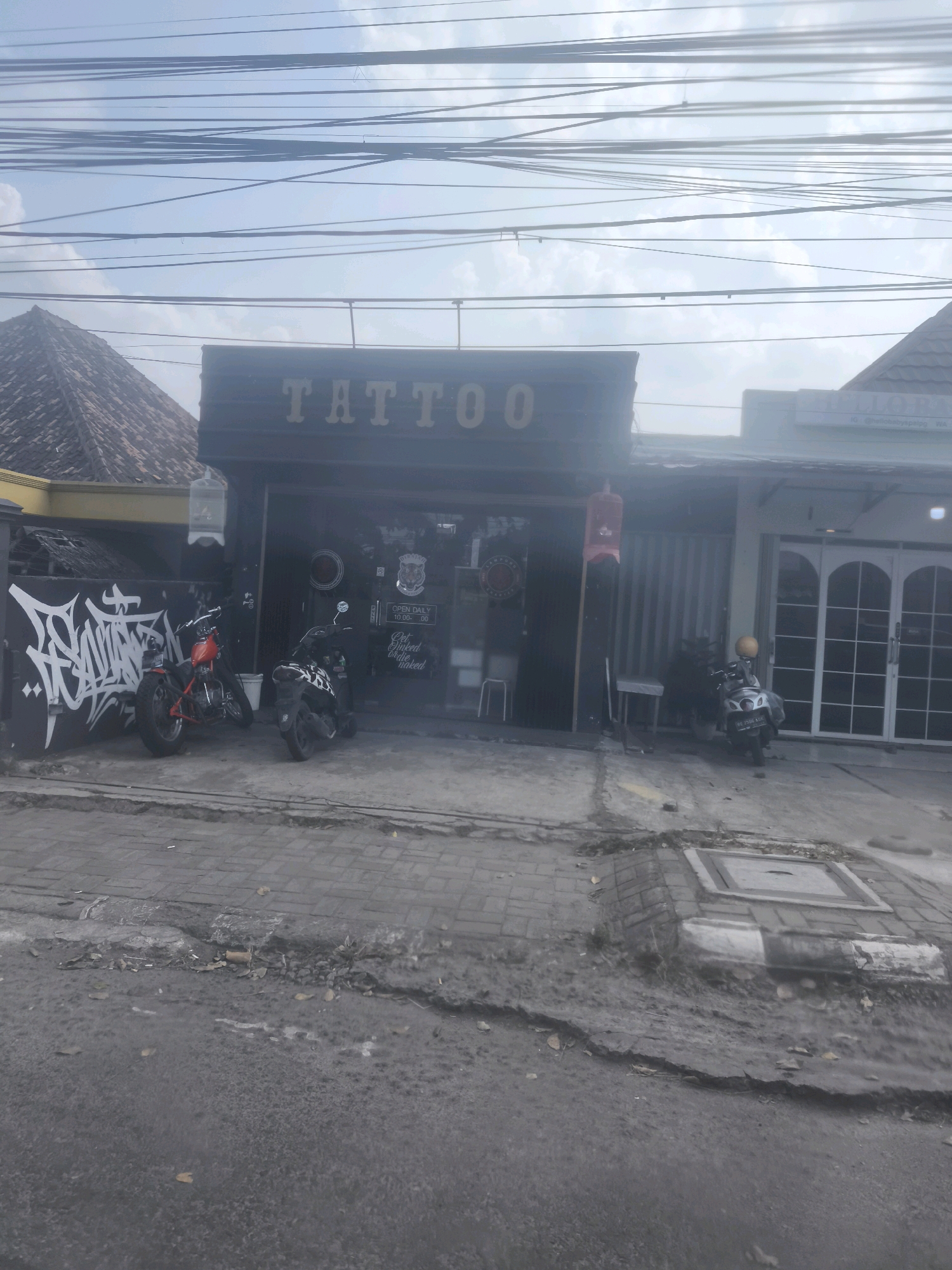 The image depicts a street view in Bandar Lampung, Indonesia, featuring a tattoo parlor with motorcycles parked in front. The tattoo parlor has a prominent "TATTOO" sign above its entrance. To the left of the entrance, there is graffiti on a black wall. Two motorcycles, one red and one black and white, are parked on the sidewalk in front of the shop. A third motorcycle, silver with a yellow helmet, is parked further down the street. The road in front of the shop appears to be paved with asphalt and brick. Overhead, a tangle of electrical wires crisscrosses the sky, a common sight in urban Indonesian landscapes. The overall impression is of a typical commercial street scene in a developing city.