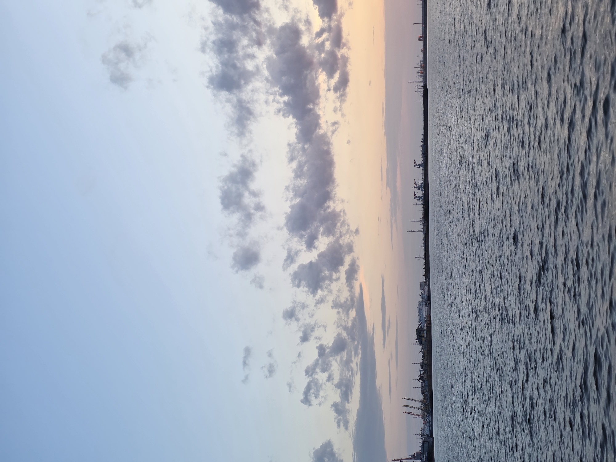 The image shows a body of water under a sky with clouds. The horizon line has some indistinct objects, potentially industrial structures or cranes, suggesting an urban or port setting. The sky transitions from a light blue on the left to a light orange hue on the right, possibly indicating sunset or sunrise. The water appears rippled, reflecting the ambient light. The image's perspective is landscape but is rotated 90 degrees clockwise. Given the city information, this scene likely depicts a view from or near the Kama River in Chaikovsky, Russia, with the distant structures possibly related to the city's industrial activities. There are no people visible in the image.