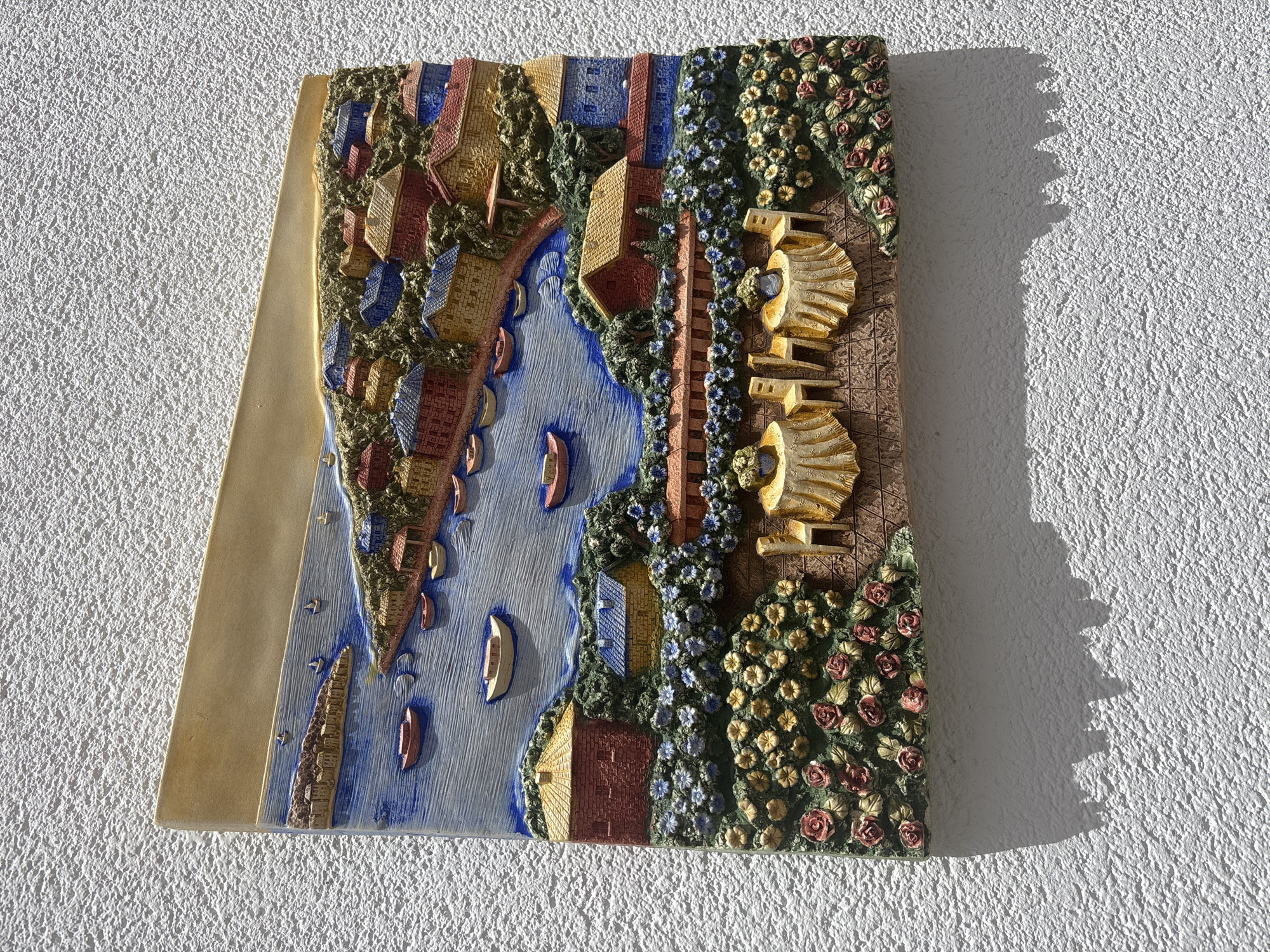 This is a detailed bas-relief sculpture depicting a coastal village scene. The sculpture is mounted on a white stucco wall, casting a shadow below it. The scene shows a body of water with several boats, a shoreline with a cluster of colorful houses with red, yellow, and blue roofs, and a hillside covered with greenery and flowers. In the foreground, there are two small tables set with chairs, each adorned with a vase of flowers, and a border of roses and other blooms. The style is somewhat whimsical and tourist-oriented, suggesting a picturesque location often found in vacation spots. While the prompt mentions Orihuela, Spain, there are no specific landmarks or identifying features within the sculpture that would definitively place it in that city. It could be a generic representation of a Mediterranean coastal town.