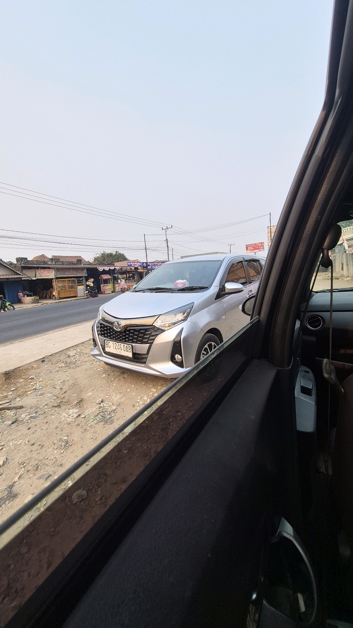A silver sedan is parked on the side of a road in Putak, Indonesia. The car, a Daihatsu Sigra based on its license plate, is facing forward with its front grille visible. The road has shops and buildings along the right side. The perspective is from inside another vehicle, looking out through the open window. The sky is clear and overcast.