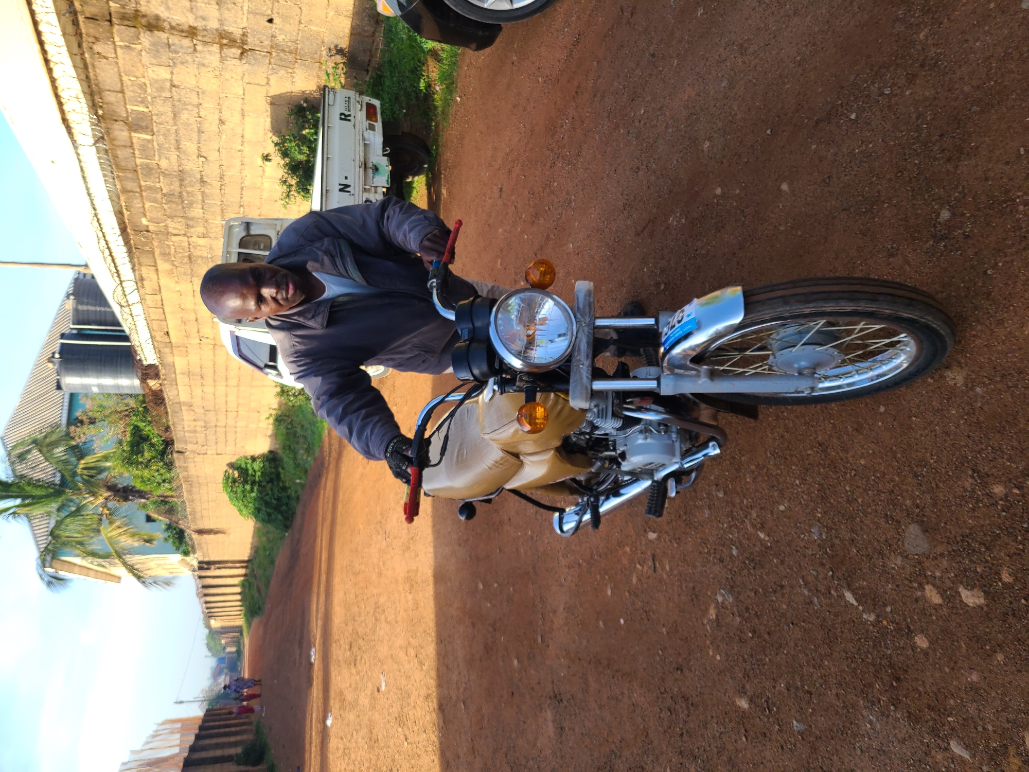 The image, set in Jos, Nigeria, captures a smiling man seated on a motorcycle on a reddish-brown dirt road. The man, with short dark hair, is wearing a dark jacket over a lighter shirt and looks directly at the camera, conveying a friendly demeanor. He is holding the handlebars of a utility-style motorcycle, which features a prominent chrome headlight, a tan or beige cushioned seat, chrome fenders, and orange turn signals.
The setting appears to be a semi-urban or rural area under a bright, clear blue sky, indicating sunny weather during the daytime. To the left, a large brick wall stretches alongside the road. Behind the man, a white pickup truck, possibly a Daihatsu Hijet as "HIJET" is visible on its side along with a partial "N-", is parked. Further in the background, multiple large black water tanks are visible, commonly used for water storage. A lush green palm tree and other vegetation are also present. The ground is unpaved, showing varying textures of dirt and small stones. On the motorcycle's front fender, a blue sticker or plate has some indistinct text. The overall scene suggests a typical roadside view in the region.
