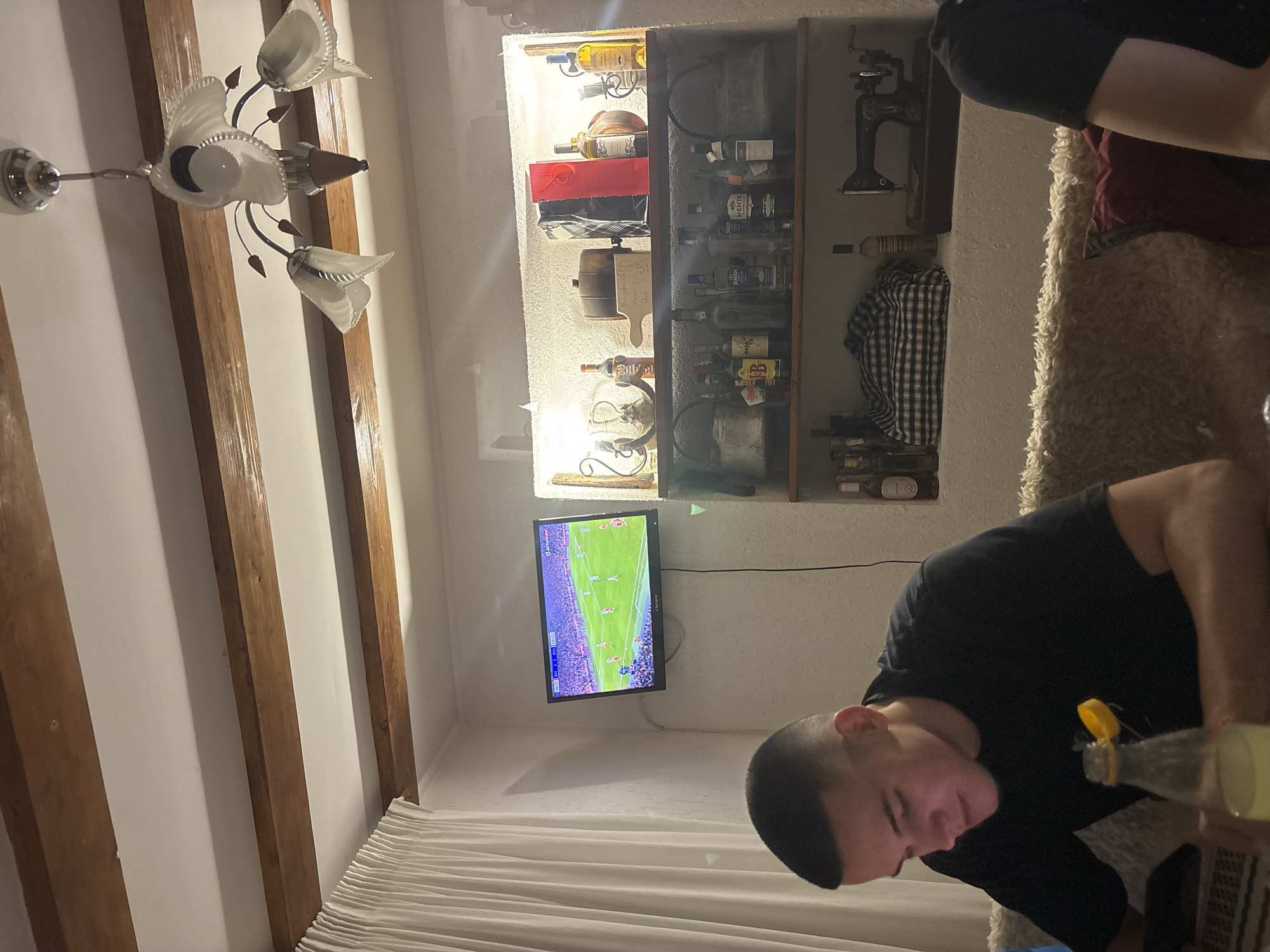 The image shows a room in Zlatograd, Bulgaria, possibly a living or entertainment space. An individual wearing a black t-shirt is centered in the right foreground, holding a bottle with a yellow cap. A television displaying a soccer game is visible on a wall. Behind the television is a recessed shelf filled with various objects, including bottles. A light fixture hangs from the ceiling on the left, with wooden beams visible along the ceiling. White curtains are seen in the lower left and middle parts of the image. An individual is sitting on a chair in the upper right.