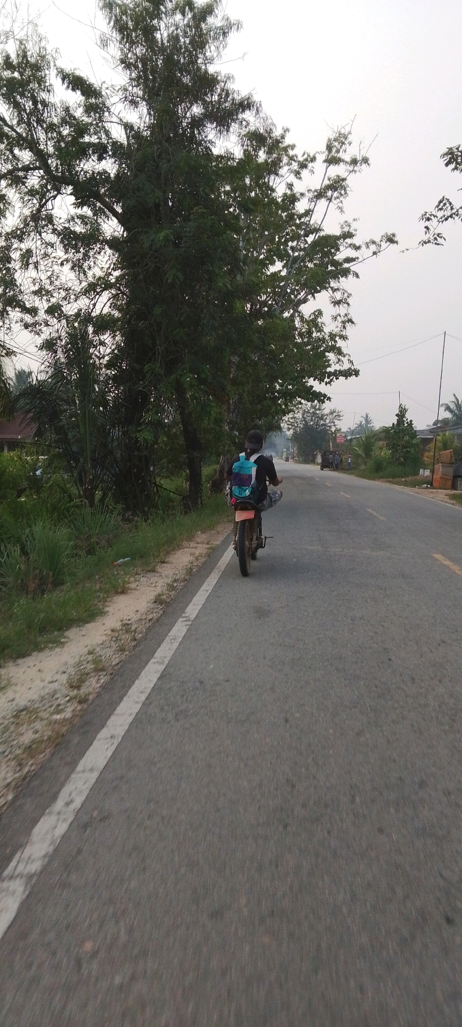 In Lenggadai Hulu, Indonesia, a person is riding a motorcycle on a paved road with a white line marking the shoulder. The rider is wearing a dark shirt and a backpack with blue, purple, and white colors. The motorcycle appears to be an older model. To the left of the road, there is a dirt shoulder with green grass and vegetation, including a large tree with dense foliage. In the background, the road continues, with other vehicles and buildings visible, suggesting a rural or suburban area. The sky is overcast.