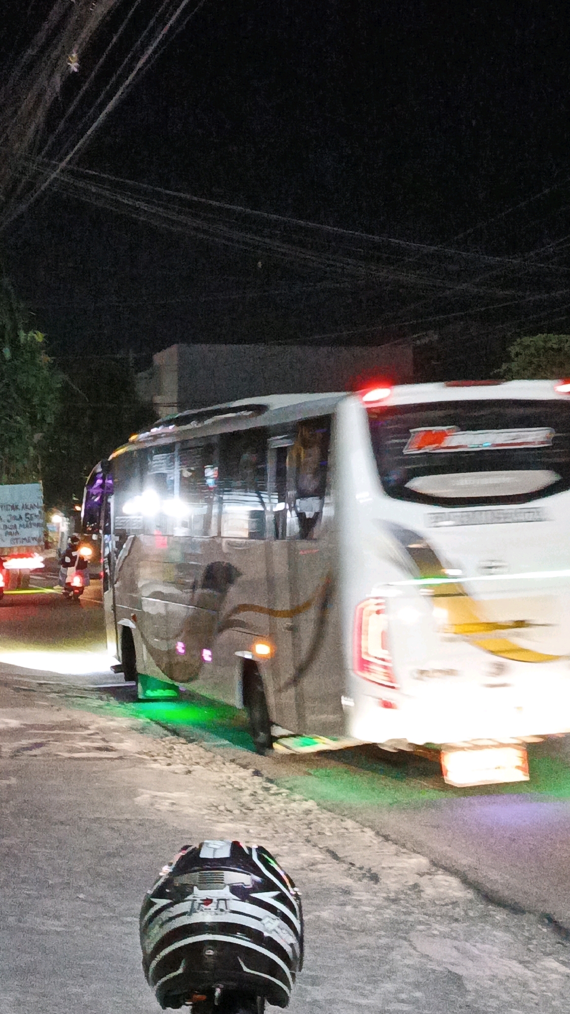 The image depicts a nighttime scene on a street in Gondangrejo, Indonesia. A white bus with tinted windows and decorative yellow and brown stripes is the primary subject, traveling from left to right and blurring due to motion. The bus has green lights illuminating its underside and red brake lights at the rear. A portion of a motorcycle helmet, black with white and silver stripes, is visible in the foreground, obscuring the lower part of the image. Other vehicles, including a truck and motorcycles, are present in the background, along with streetlights and overhead wires, indicating an urban environment. The ground appears to be asphalt, with some gravel on the side of the road. A building is visible in the far background.