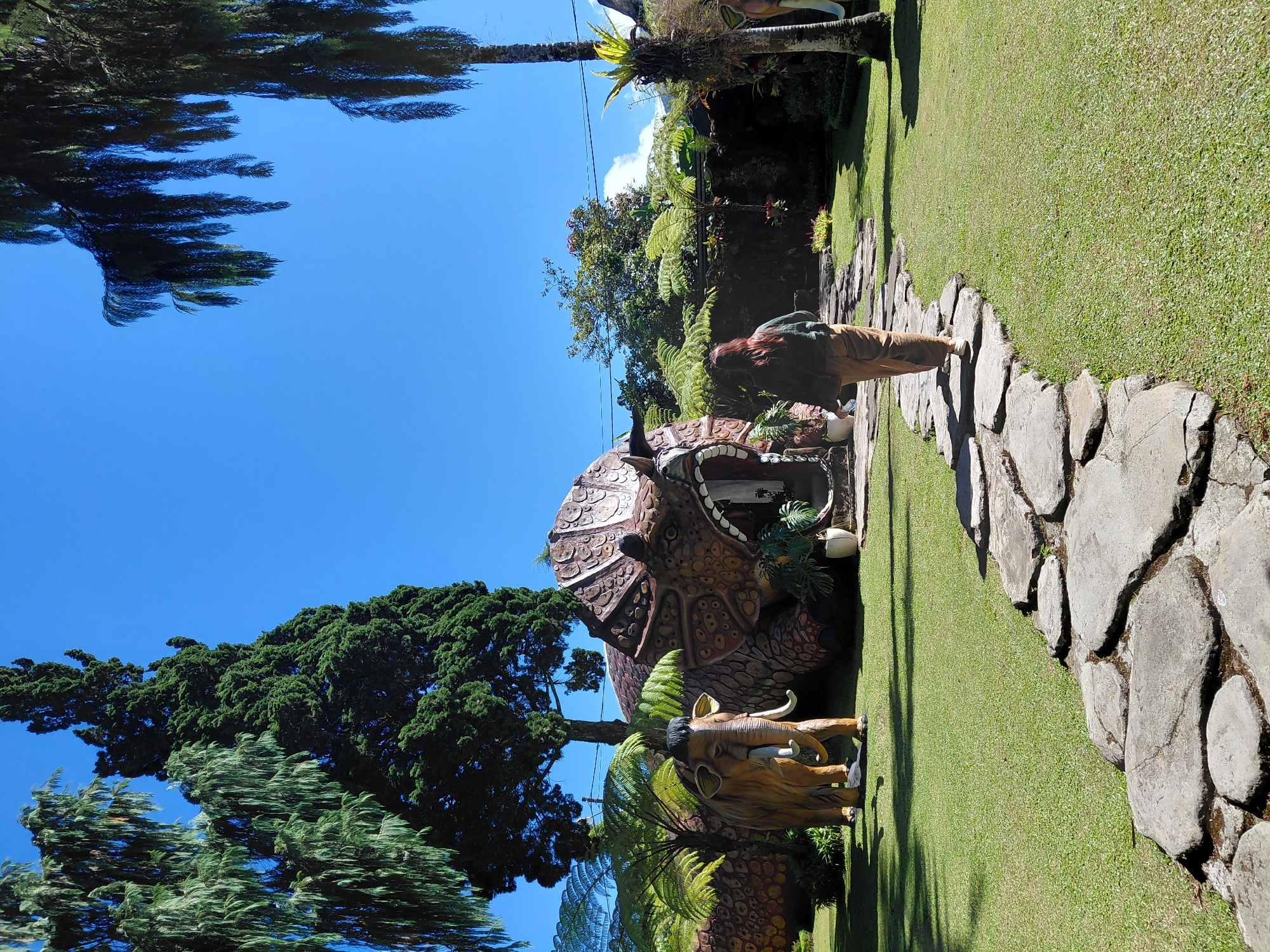 A person walks on a stone pathway through a grassy area. To the left of the path is a large, dinosaur-shaped building with an open entrance. A statue of a mammoth stands to the left of the dinosaur building. To the right of the path, palm trees and other vegetation are visible. The sky is clear and blue. The scene appears to be a theme park or attraction. The location is identified as Bali, Indonesia.