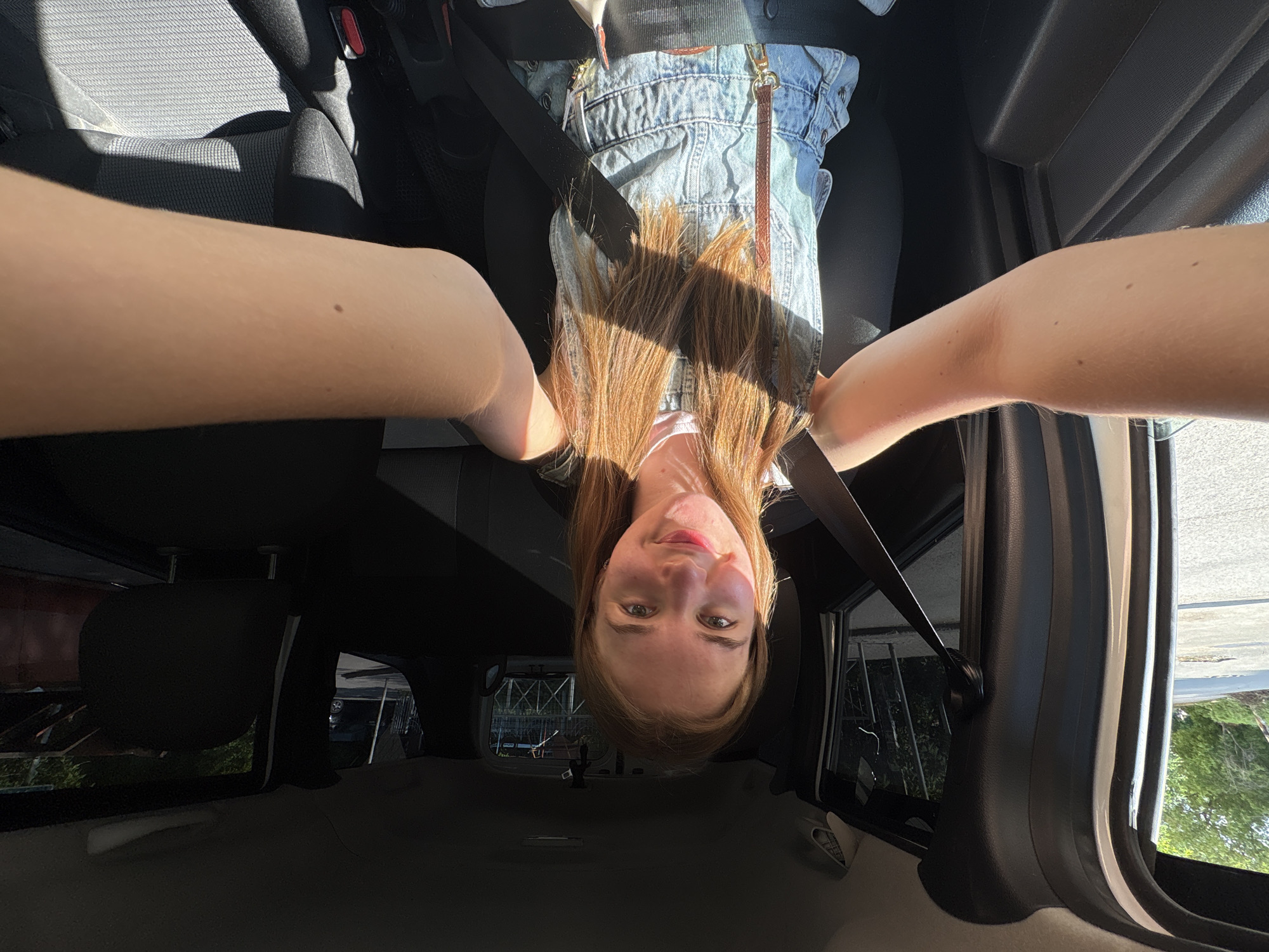 A selfie-style, upside-down image shows a woman in the driver's seat of a car. She is wearing denim overalls and a seatbelt. Her reddish-brown hair hangs down in front of her. The car's interior is visible, including the seats and windows. The background shows an exterior scene with a metal structure and greenery, likely indicating an urban area. There is no available information to confirm the location of the image to be in Perm, Russia.