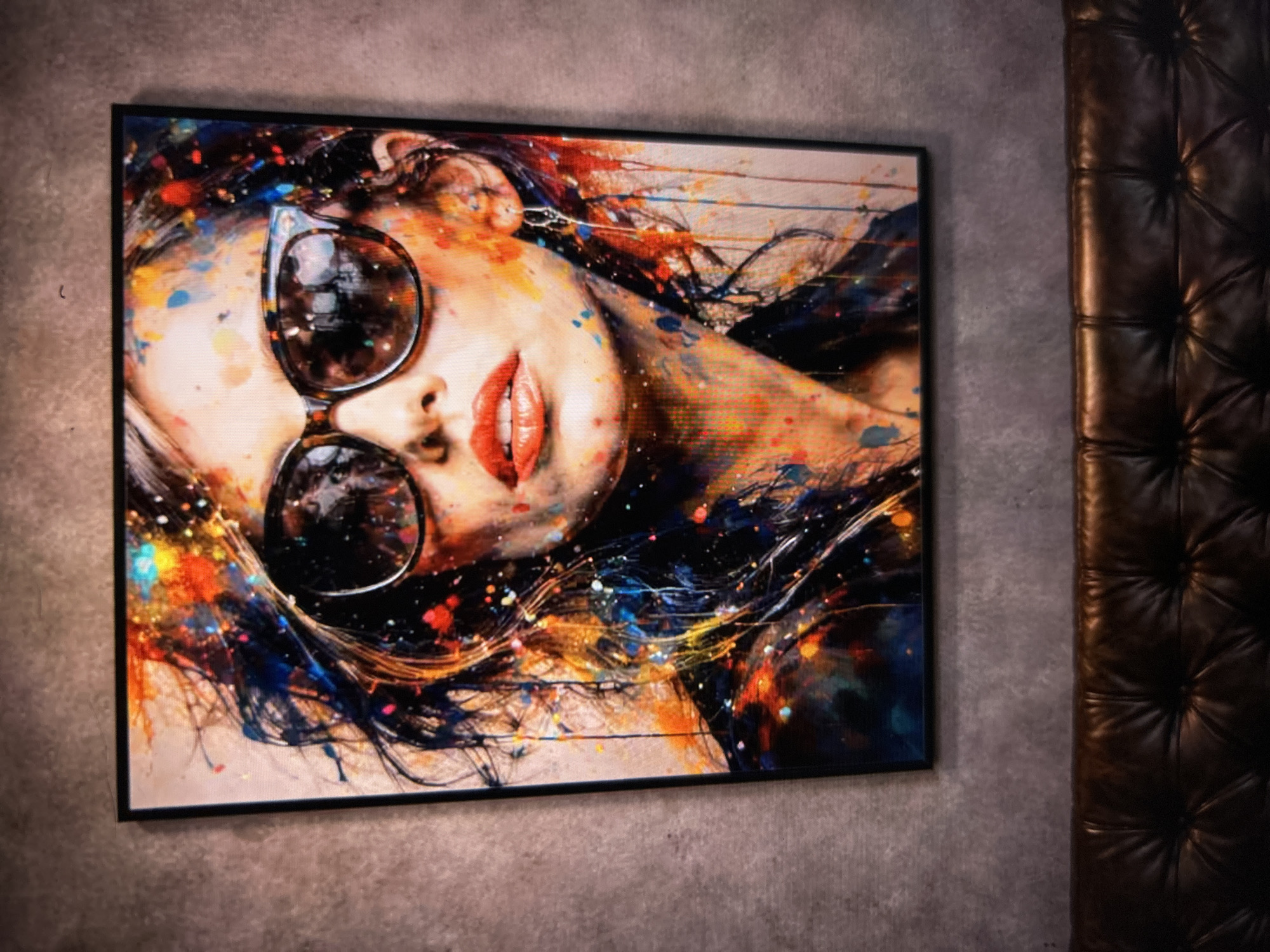 A framed painting of a woman with sunglasses and red lipstick hangs on a textured gray wall above a dark brown leather couch. The painting features a vibrant, splattered paint style, with bold colors of orange, yellow, blue, and red across the woman's face and hair. Her eyes are obscured by large, tortoise-shell sunglasses. The couch is tufted and appears to be made of leather. The overall scene suggests an indoor setting, likely a living room or lounge area. The city of "Unknown, Unknown" is not depicted in the image.