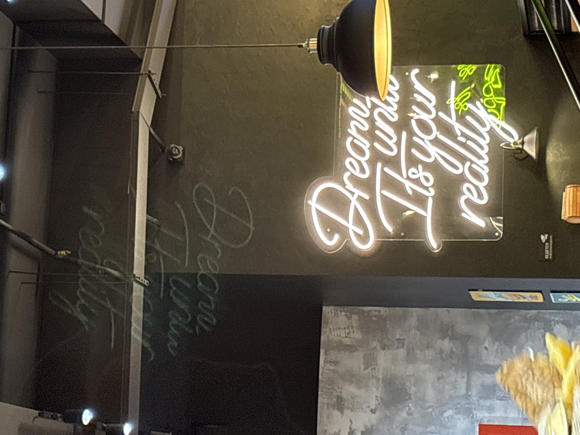 This is an indoor shot of what appears to be a modern cafe, bar, or restaurant in Izhevsk, Russia. The primary subject is a white neon sign mounted on a dark green-grey wall. Above the sign, a black pendant light with a yellow interior hangs from the ceiling. To the left, an industrial-style pipe or beam structure is visible, along with reflections of light on the dark wall, which also show a faint, chalk-like rendering of parts of the neon sign's text.
The white neon sign clearly displays the motivational phrase: "Dream until It's your reality." Above the word "reality," there is additional smaller text in green neon, rendered in a different font, which is difficult to read definitively but appears to be a short, complementary phrase, possibly "Live Your Story" or "Live Your Life."
Below the main wall, there is a dark shelf or counter, and further down, a textured grey wall. To the far right, a small wall-mounted gold-colored spotlight is visible, along with a light-colored wooden decorative object and part of a yellow, dried plant arrangement. The artificial lighting, dominated by the neon sign and pendant lamp, suggests an indoor evening setting or an establishment designed with atmospheric mood lighting. No people are present in the image.