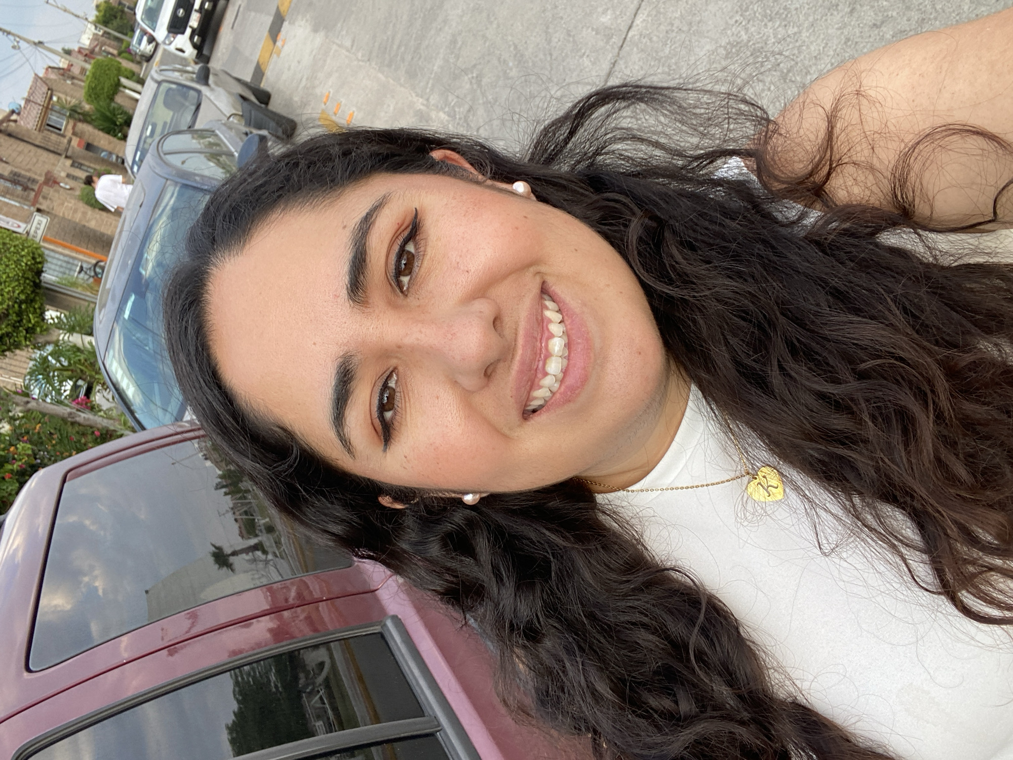 A woman with dark, wavy hair, brown eyes, and a white t-shirt smiles at the camera. She wears pearl earrings and a gold necklace with a heart-shaped pendant. A burgundy vehicle is partially visible in the background. Other cars and buildings are seen in the distance. The photo appears to have been taken in San Bernardino Tlaxcalancingo, Mexico.