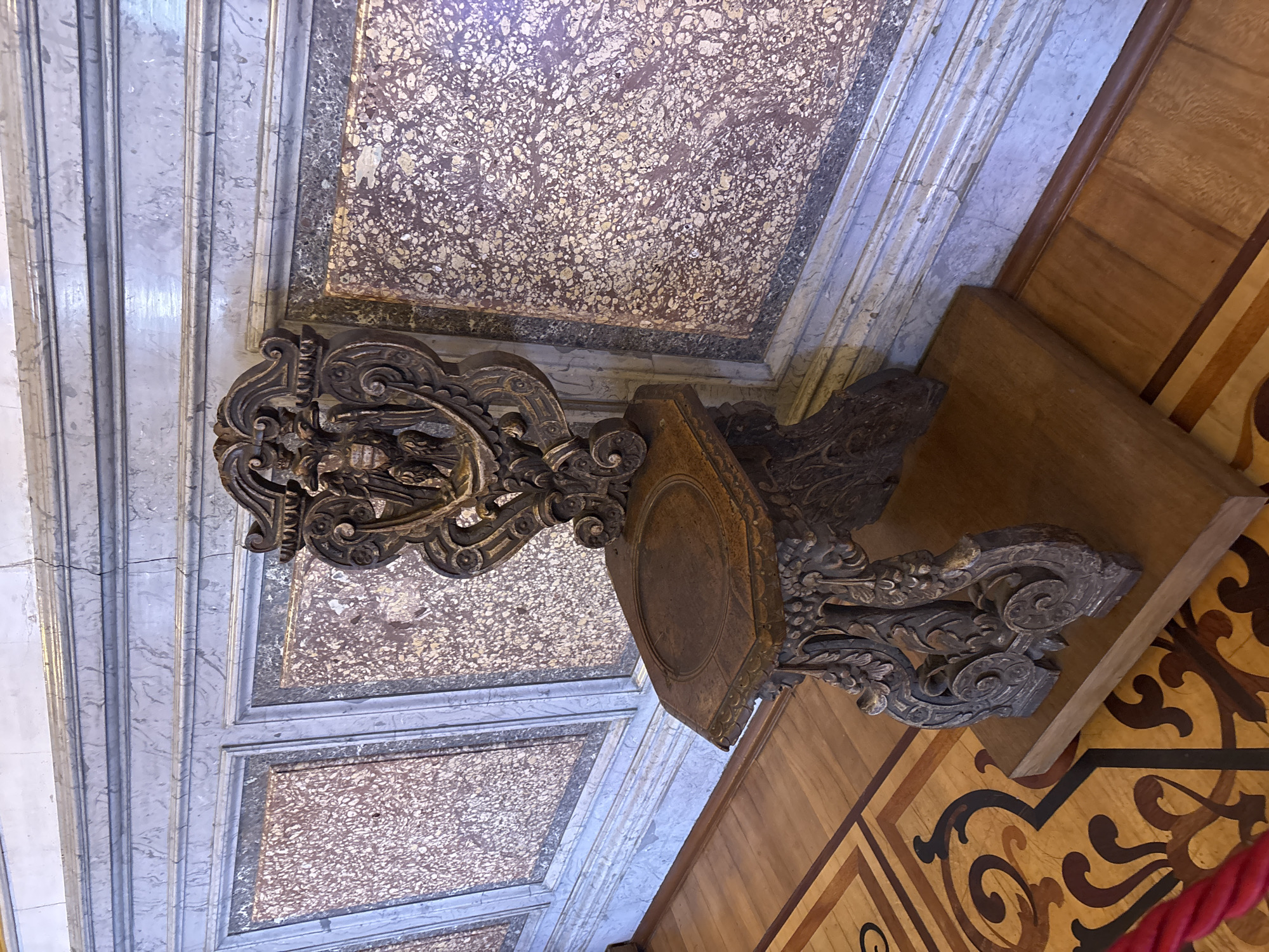 The image shows a highly ornate, dark wood chair with intricate carvings, including what appears to be an eagle or griffon motif on the backrest. The chair is positioned on a wooden platform in front of a wall paneled with marble and a decorative border. The floor is made of polished parquet with an elaborate inlaid pattern. The overall setting suggests a historical or museum interior. This chair is located in the Catherine Palace in the city of Pushkin, near Saint Petersburg, Russia, specifically in the Amber Room or one of its antechambers, which are part of the palace complex. There are no people visible in the image.