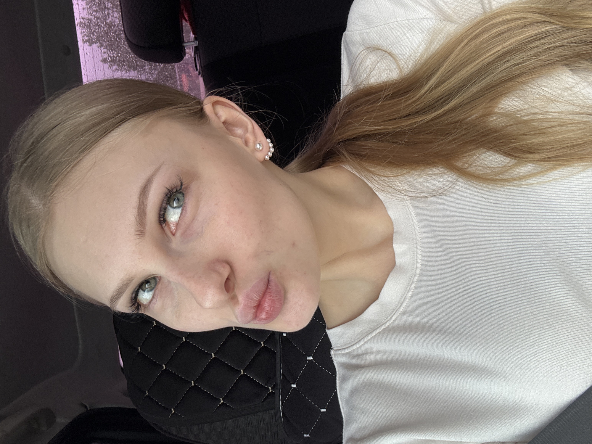A young woman is pictured in an intimate, slightly angled portrait, likely taken indoors with a dark, blurred background. The woman has fair skin, blue-green eyes, and light blonde hair pulled back into a ponytail. She has fair skin with some blemishes. She is wearing a white t-shirt and small pearl earrings. She is wearing a black quilted mask. The image could be situated in Perm, Russia.
