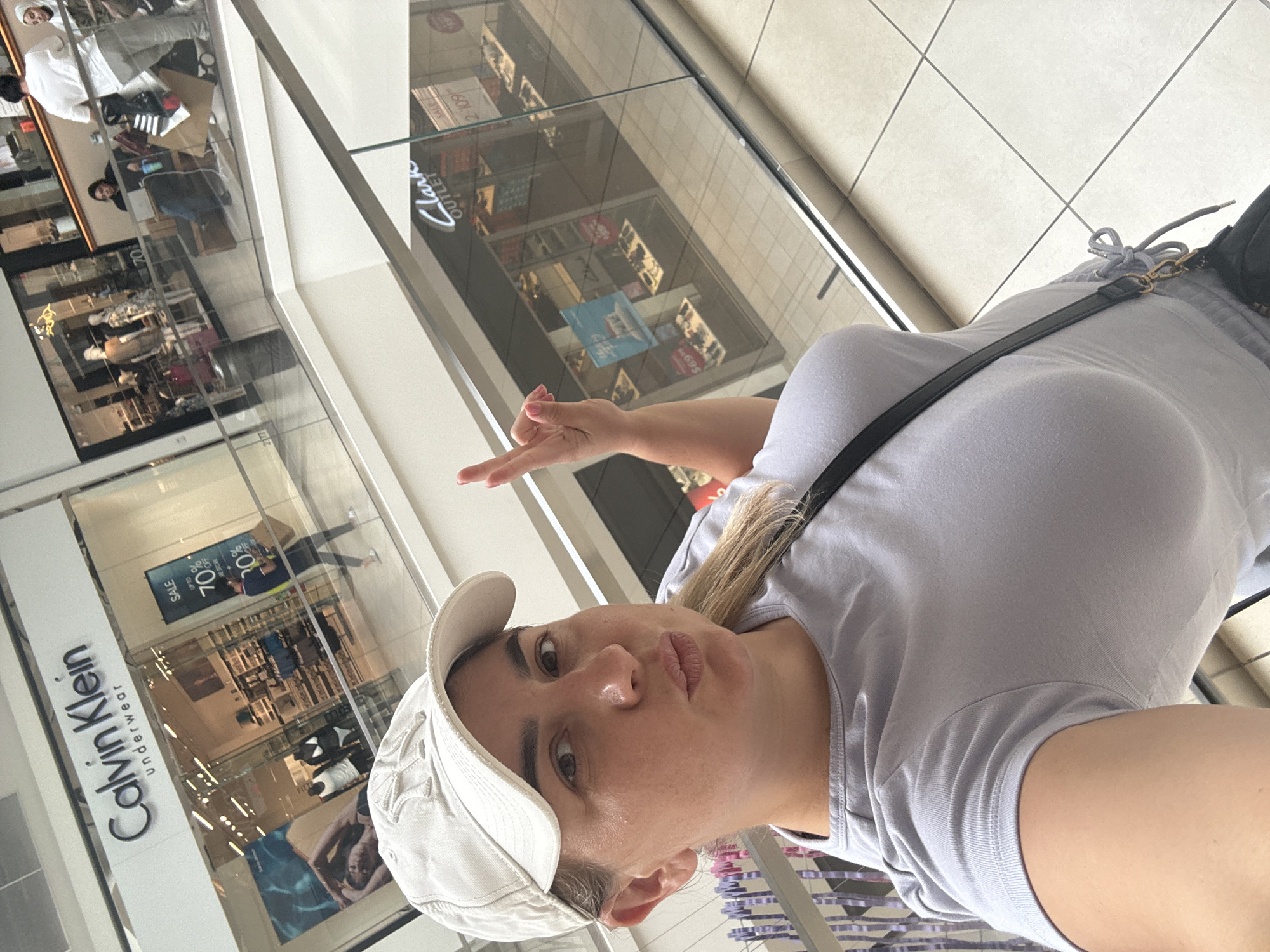 A woman wearing a light grey t-shirt and a white baseball cap poses for a selfie in a shopping mall. The background shows a Calvin Klein underwear store with a "SALE UP TO 70% OFF" sign. Another store, "Clark's Outlet," is visible across the mall, with shoes displayed in its window. The woman is holding her left hand up, making a peace sign with her index and middle fingers. She appears to be in a shopping mall in Rosemont, United States of America, likely in the Fashion Outlets of Chicago.