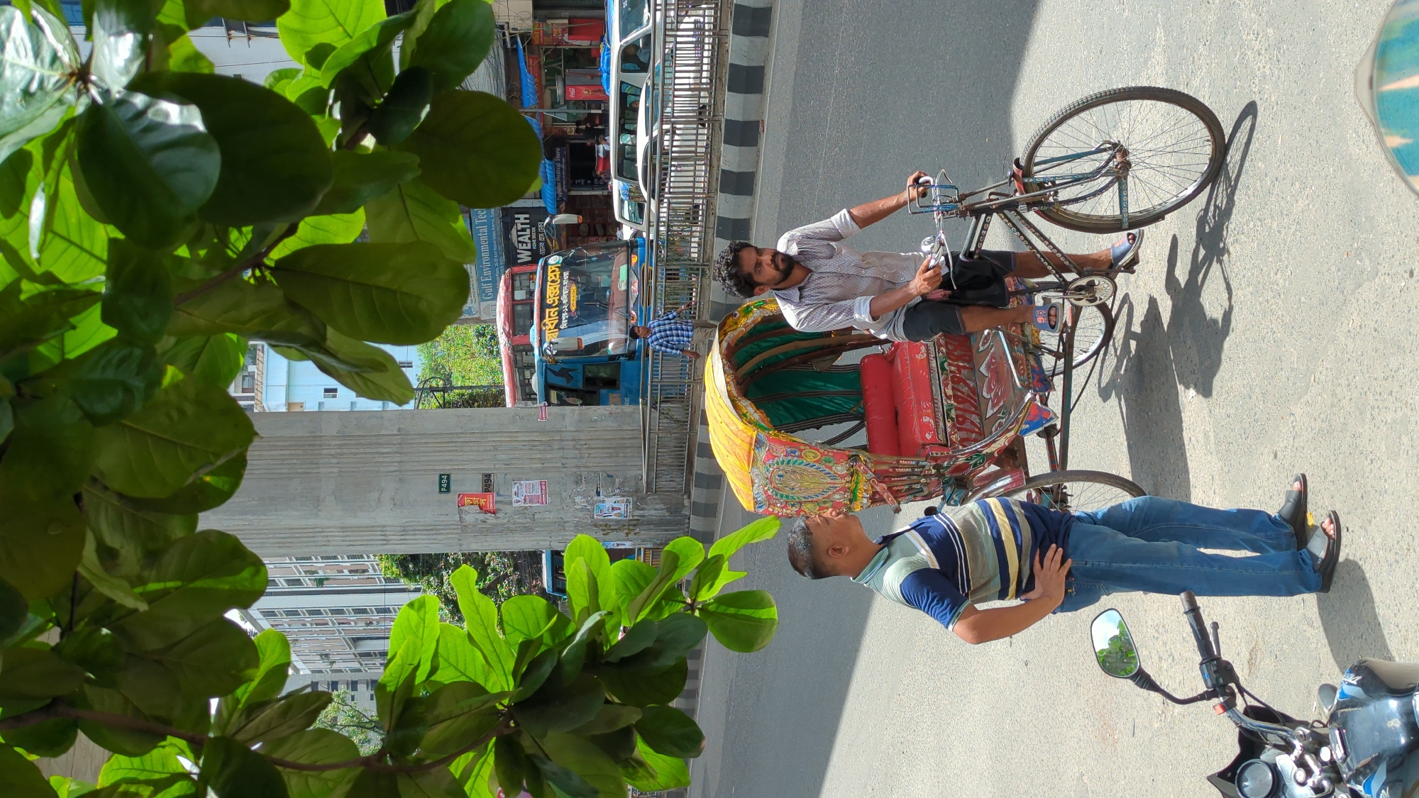 In Dhaka, Bangladesh, a rickshaw driver cycles a brightly colored, ornate rickshaw on a street. He is wearing a patterned short-sleeved shirt and dark shorts. Beside the rickshaw, a man in a striped polo shirt and blue jeans stands with his hands on his hips, looking to the left. In the background, a blue bus with Bengali text on its windshield is stopped, and behind it, other vehicles and buildings are visible. Large green leaves from a tree hang over the scene, casting shadows on the road. A motorcycle is partially visible in the lower left corner. The scene depicts a typical urban streetscape in Dhaka, with its vibrant rickshaws and bustling traffic.