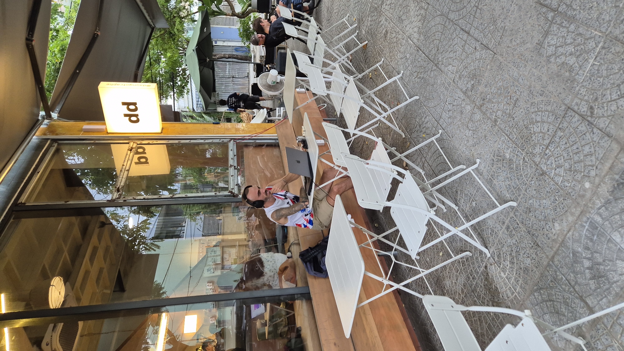 An outdoor seating area of a cafe in Da Nang, Vietnam, features white metal tables and chairs arranged on a patterned tiled floor. A man with headphones sits at a table, looking at a laptop, with a backpack next to him. In the background, other patrons are seated at tables, and a fan is visible. A lighted sign with "dd" is mounted on the building's exterior, reflecting in the glass storefront. Trees and awnings provide shade.