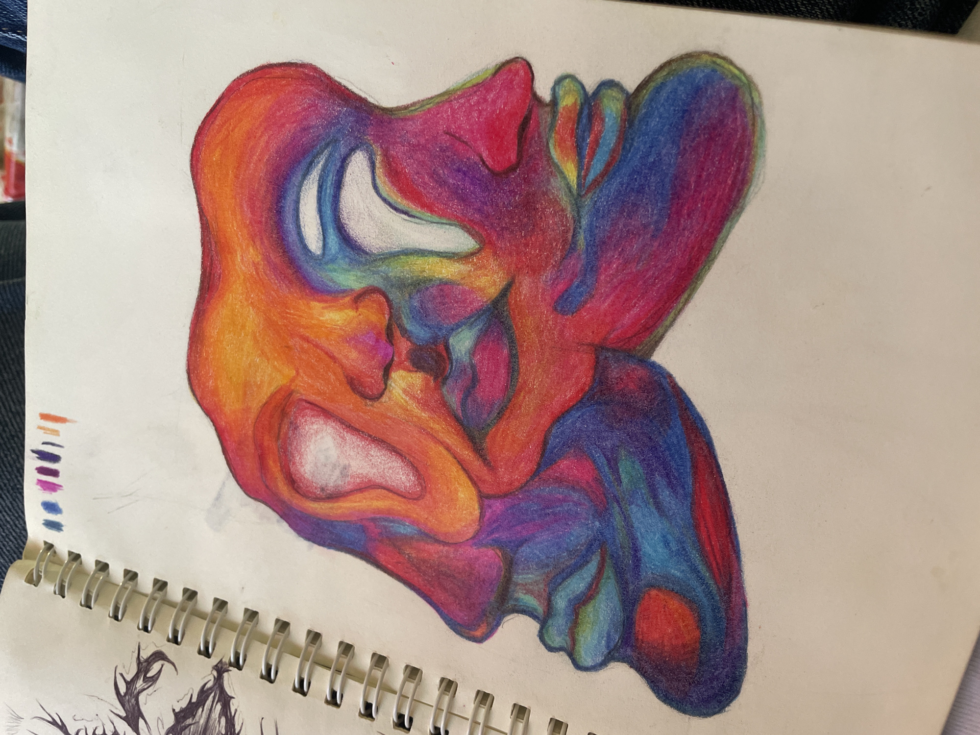 The image shows a colored pencil drawing on a page in a spiral-bound sketchbook. The drawing is an abstract, organic shape filled with blended colors of orange, red, purple, and blue. The shape appears to be composed of interconnected, rounded forms with lighter, white or pale-colored areas within. The drawing is positioned in the center of the page, with parts of other drawings visible on adjacent pages. There is no location given in the image.