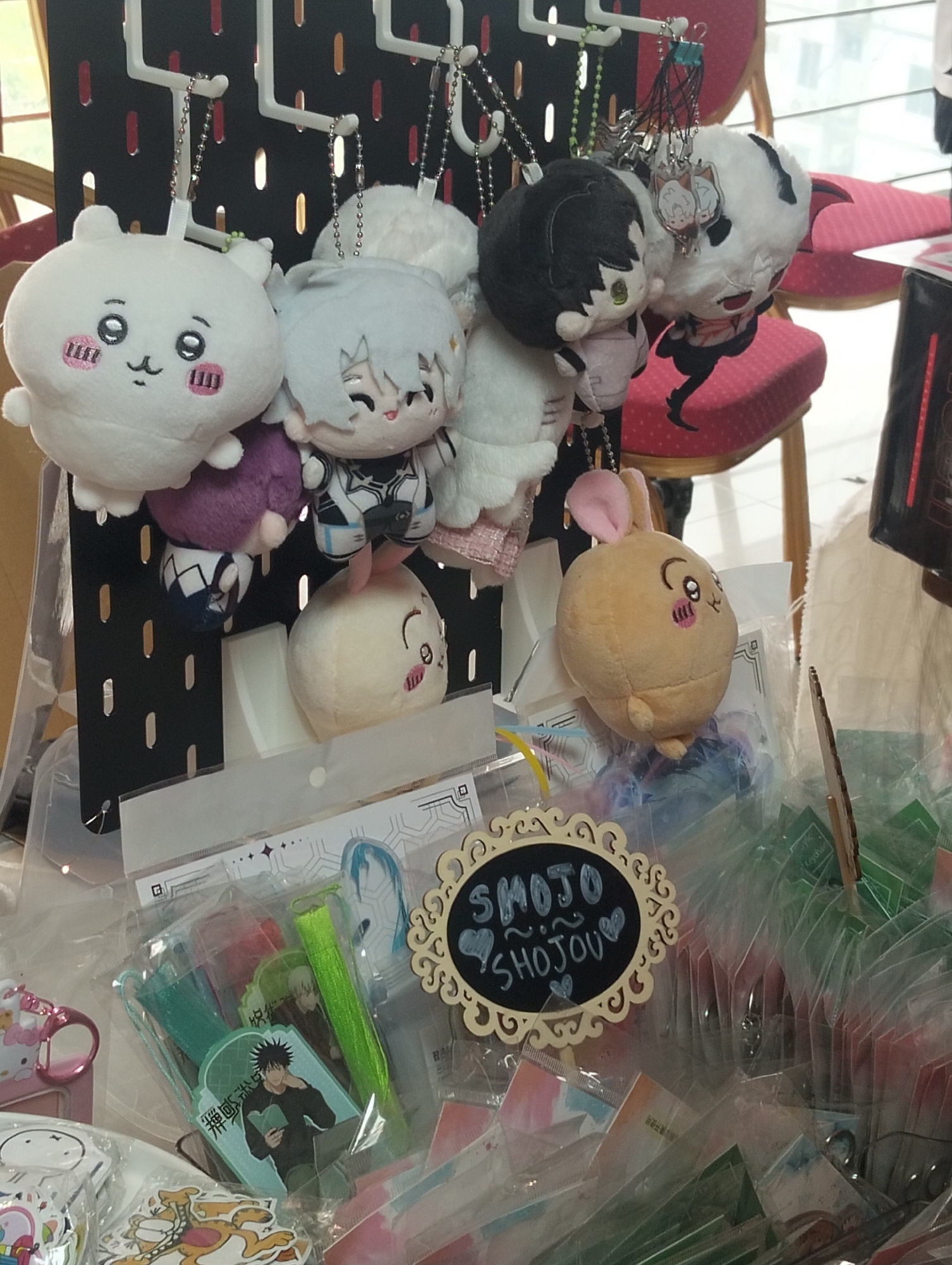 The image shows a close-up of various anime-themed merchandise displayed on a black pegboard and in plastic packaging. In the foreground, there are several plush keychains of anime characters hanging from the pegboard. One is a white round plush with pink cheeks and a smiling face. Next to it is a light blue-haired anime character plush. To the right, there's a black-haired anime character plush and a white and black plush with pointed ears. Further to the right, a light brown round plush with pink ears and a closed-eyed smiling face is visible.
Below the hanging keychains, plastic-wrapped items are displayed. These include smaller plush keychains, stickers, and what appear to be trading cards or decorative inserts featuring anime characters. One prominently displayed item in the lower middle is a set of green ribbons and what looks like an acrylic standee with an anime character reading a book.
To the right of these items, there is a decorative wooden sign with the words "SMOJO SHOJOU" written in chalk, surrounded by decorative swirls and small heart shapes. Behind this sign, there are numerous small plastic-wrapped items, mostly in shades of green and red.
The background is blurred, but it suggests an indoor environment, possibly a market stall or a shop. The lighting is neutral, illuminating the merchandise clearly. There are no people visible in the frame. The overall scene depicts a retail display of popular culture merchandise.