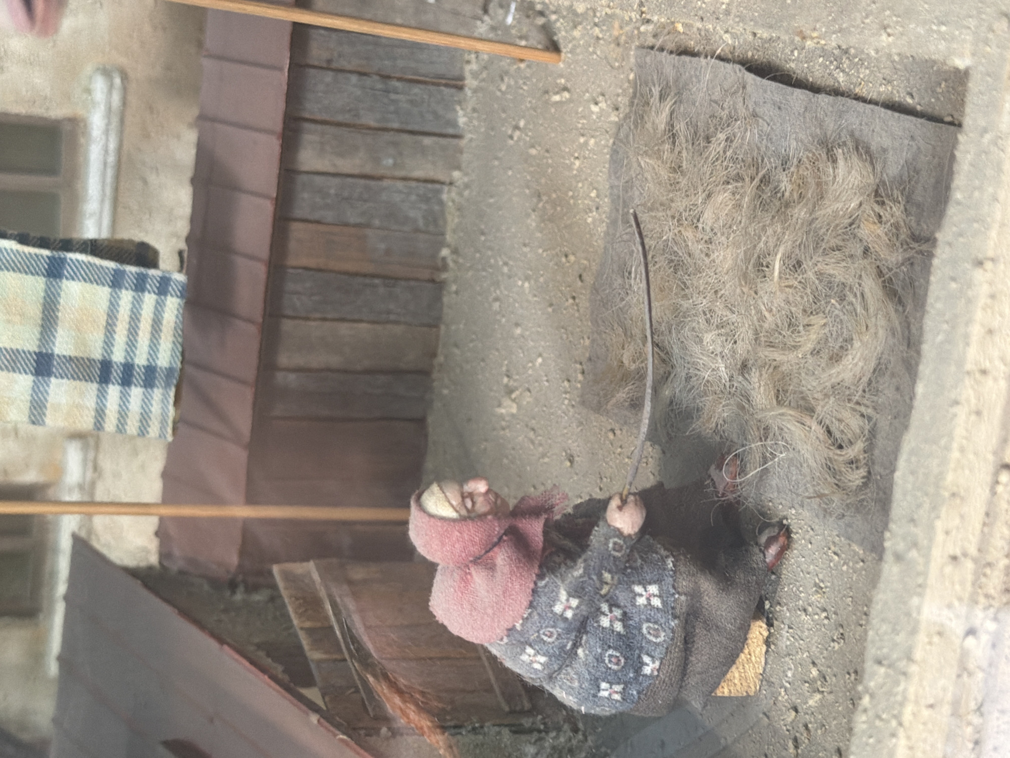 The image shows a miniature diorama depicting a scene in Tbilisi, Georgia. In the foreground, a doll-sized elderly woman with a hunched back and a pink headscarf is seated on the ground. She is wearing a dark skirt and a patterned sweater with white and grey floral designs. Her right hand holds a thin stick or tool, which she appears to be using to work with a pile of fibrous material, possibly straw or hair, spread out on a dark fabric mat on the ground. The ground is covered in dirt and small pebbles. Behind the woman, a rustic wooden fence or wall is visible, with a sloped, reddish-brown roof extending from a building to the left. A checkered blanket in blue and white is draped over what appears to be a clothesline or a beam. The overall environment suggests an outdoor setting, possibly a yard or alleyway, characteristic of older urban areas in Tbilisi. The diorama is detailed, aiming to recreate a realistic depiction of a historical or traditional scene.