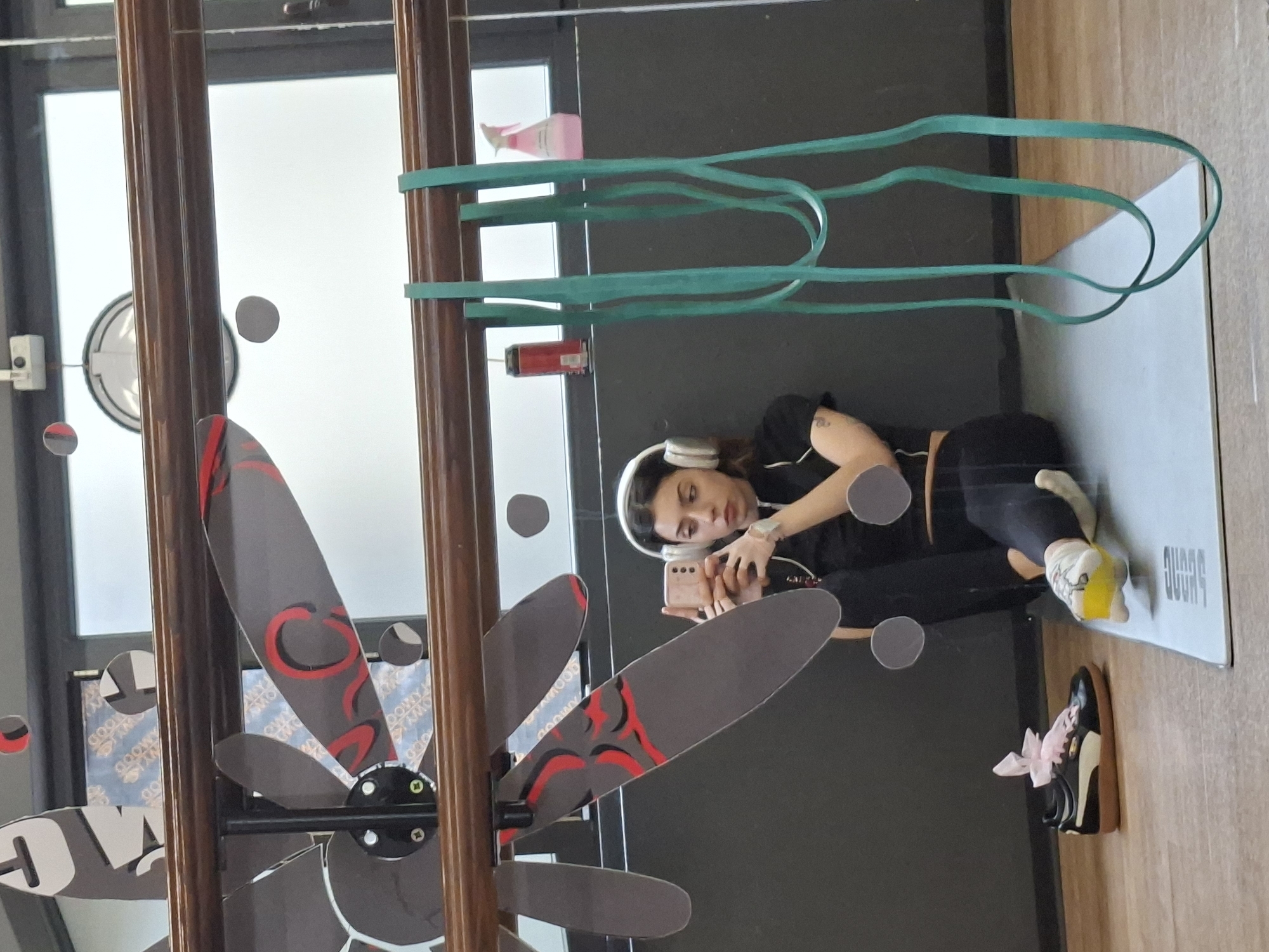 The image captures a young woman lying on a light grey yoga mat on a wooden floor, appearing to take a selfie or record herself with a light-colored smartphone. She wears white over-ear headphones and dark athletic wear, consisting of a short-sleeved top and leggings. A tattoo is visible on her left arm.
The setting appears to be an indoor space, possibly a gym, yoga studio, or versatile common area, indicated by the fitness equipment and large windows letting in natural light. Several tall, dark wooden poles or frames run vertically across the frame, partially obstructing the view.
To the right of the woman, green resistance bands are draped over one of these wooden structures. A pink spray bottle and a red and white can (possibly a beverage) are also visible near these bands. On the wooden floor near her feet is a pair of black shoes with white soles and a decorative pink bow.
To the left of the woman, a distinctive decorative object resembling a multi-bladed fan or abstract sculpture is attached to another wooden pole. Its grey blades are adorned with red, swirling designs. Through the gaps in this object, patterned text that appears to be reflections, possibly reading "OWMOO" or "GODNY" in reverse, can be vaguely discerned.
The yoga mat has the word "OVWNZ" or "OVWNS" printed in white text. The scene is brightly lit, suggesting it is daytime. The woman's expression is neutral and focused on her phone.