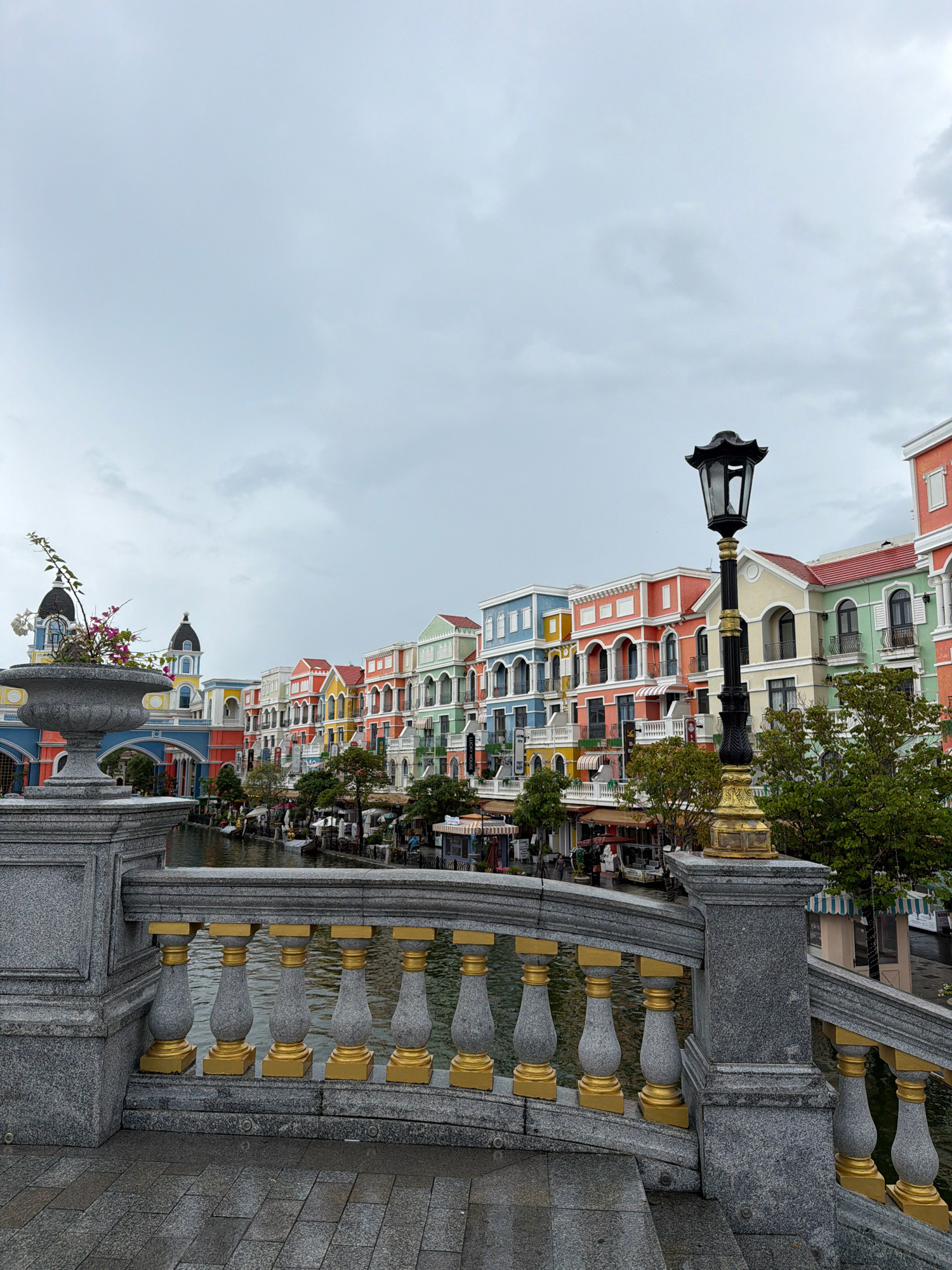 This is a full-shot, eye-level, outdoor photograph of a picturesque canal lined with colorful buildings in the city of Phú Quốc, Vietnam. The buildings are styled with Mediterranean architecture, featuring a variety of pastel colors such as blue, yellow, orange, pink, and green. They have white trim, arched windows and balconies with black railings, and red tiled roofs. The canal reflects the cloudy, overcast sky.
In the foreground, a stone bridge railing with ornate gold accents and decorative balusters overlooks the canal. To the left, a large stone planter holds a small plant with pink flowers and green leaves. A lamppost with a black, oriental-style top and a gold base stands prominently on the bridge. The ground on the bridge is paved with grey stone tiles.
Along the canal, there are trees and awnings of shops and restaurants. A few small boats are visible in the water. The sky is a uniform, dull gray, suggesting a cloudy day. The overall scene evokes a European-style waterfront promenade, likely within a resort or themed area in Phú Quốc.