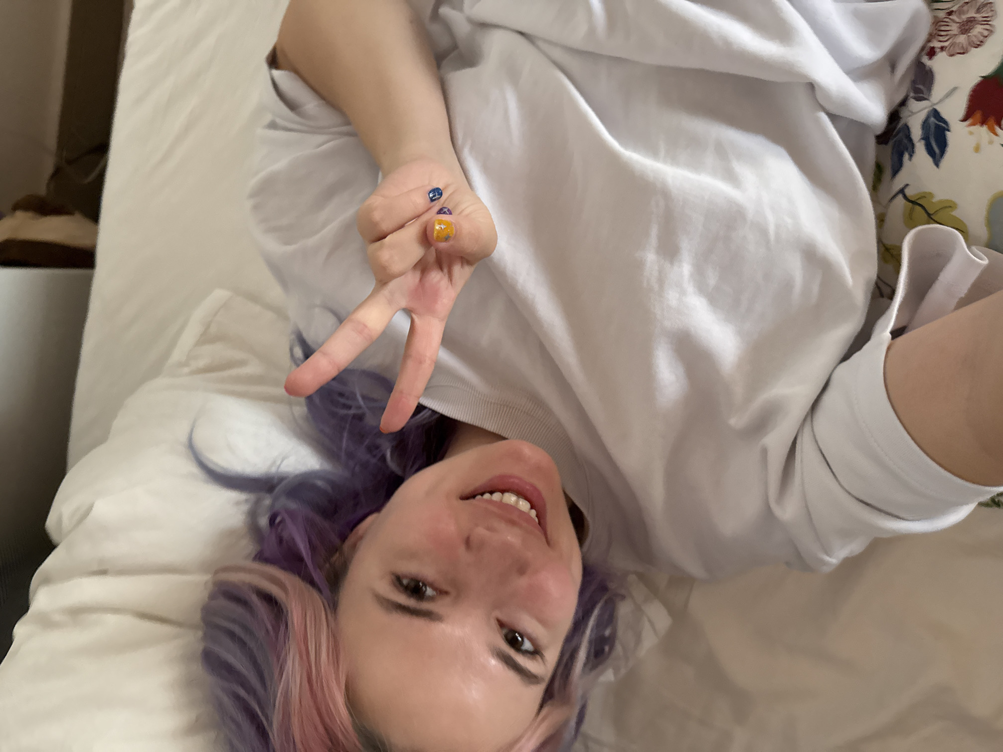 A young woman is lying on a white bed. She is wearing a white sweatshirt and has dyed purple and pink hair. She is smiling and holding up a peace sign with her right hand. Her fingernails are painted in different colors and patterns. A floral pillow is visible behind her right shoulder. A bedside table with an object on it is visible in the background. The image does not provide sufficient details to verify a specific location in Zagreb, Croatia.