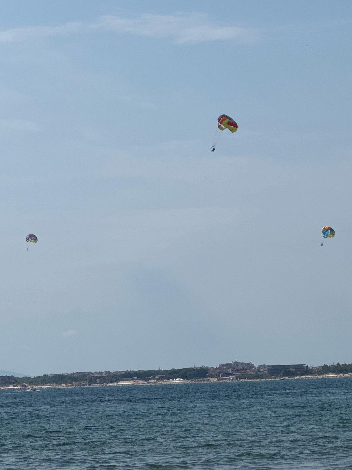 The image depicts a wide, tranquil blue sea in the foreground with gentle waves under a hazy sky. In the middle ground, a long, narrow coastline is visible, lined with buildings and lush green trees, indicating a developed area. The cityscape appears to be the historic old town of Nesebar, Bulgaria, situated on a peninsula. Three colorful parasails are suspended high in the sky, each carrying a person. One parasail, predominantly red and yellow, is positioned towards the upper left of the frame. Another, a vibrant mix of green, yellow, and blue, floats closer to the center. The third parasail, a multi-colored design with red, blue, and yellow accents, is located on the far right. The parasails are tethered to unseen boats or a towing vehicle, suggesting recreational activity. The overall scene captures a serene summer day at a coastal resort.
