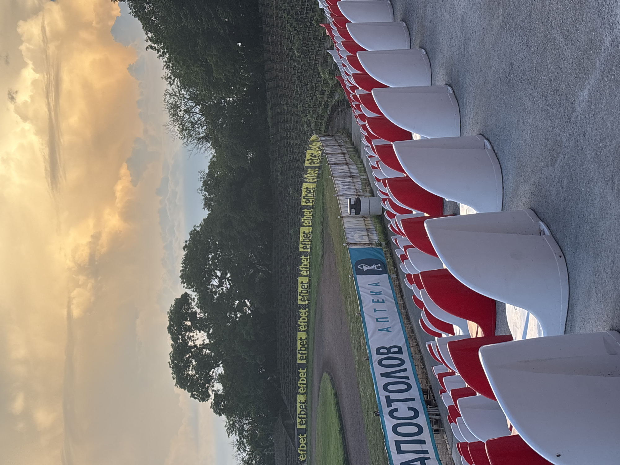 This media file captures an empty outdoor sports stadium or track and field facility in Kazanlak, Bulgaria, during the golden hour. The scene is dominated by rows of red and white stadium seats, clean and unoccupied, curving along the right side of the frame. Adjacent to the seating area is a dirt track, partially encircling an inner grassy field.
Dense green trees form a backdrop to the left of the track, their foliage reaching towards a dramatic sky filled with large, warm-toned clouds. These clouds are illuminated by the soft, golden light of what appears to be a setting or rising sun, creating a tranquil atmosphere. Patches of lighter blue sky are visible between the cloud formations.
No people or activities are taking place, leaving the stadium in a quiet state. Visible text includes a long yellow banner running along the track, repeatedly displaying "efbet," which is a Bulgarian betting company. Another prominent blue and white banner, partially visible in the foreground, features the text "АПОСТОЛОВ АПТЕКА" (Apostolov Pharmacy) along with a pharmacy cross logo, providing clear location cues for Bulgaria. The overall impression is one of peaceful solitude at the sports venue.