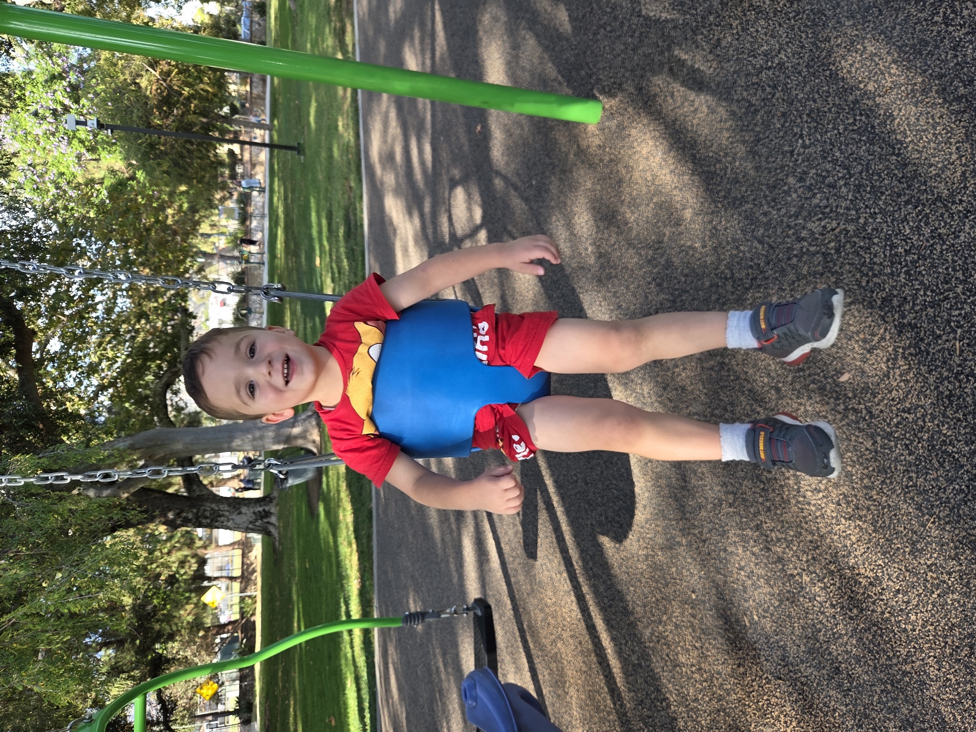 A young boy is seated in a swing at a park. The boy is wearing a red t-shirt with a yellow cartoon character on it, red shorts with white lettering, white socks, and gray sneakers with red accents. He is in a blue swing seat that supports his torso. The swing is attached to metal chains that are suspended from a green metal frame. The boy is smiling and looking towards the camera.
The park appears to be located in Glendale, California, United States of America. The ground is covered in a textured, dark brown surface, likely a safety material common in playgrounds. Shadows of trees and playground equipment are cast across the ground, indicating an outdoor setting during the daytime. In the background, there are trees with green foliage, and some grass. There are also other playground structures and a pathway visible in the distance, along with people walking. A blue plastic seat is visible to the left of the boy, possibly another part of the swing set or a different piece of playground equipment. The overall scene depicts a child enjoying time at a public park.