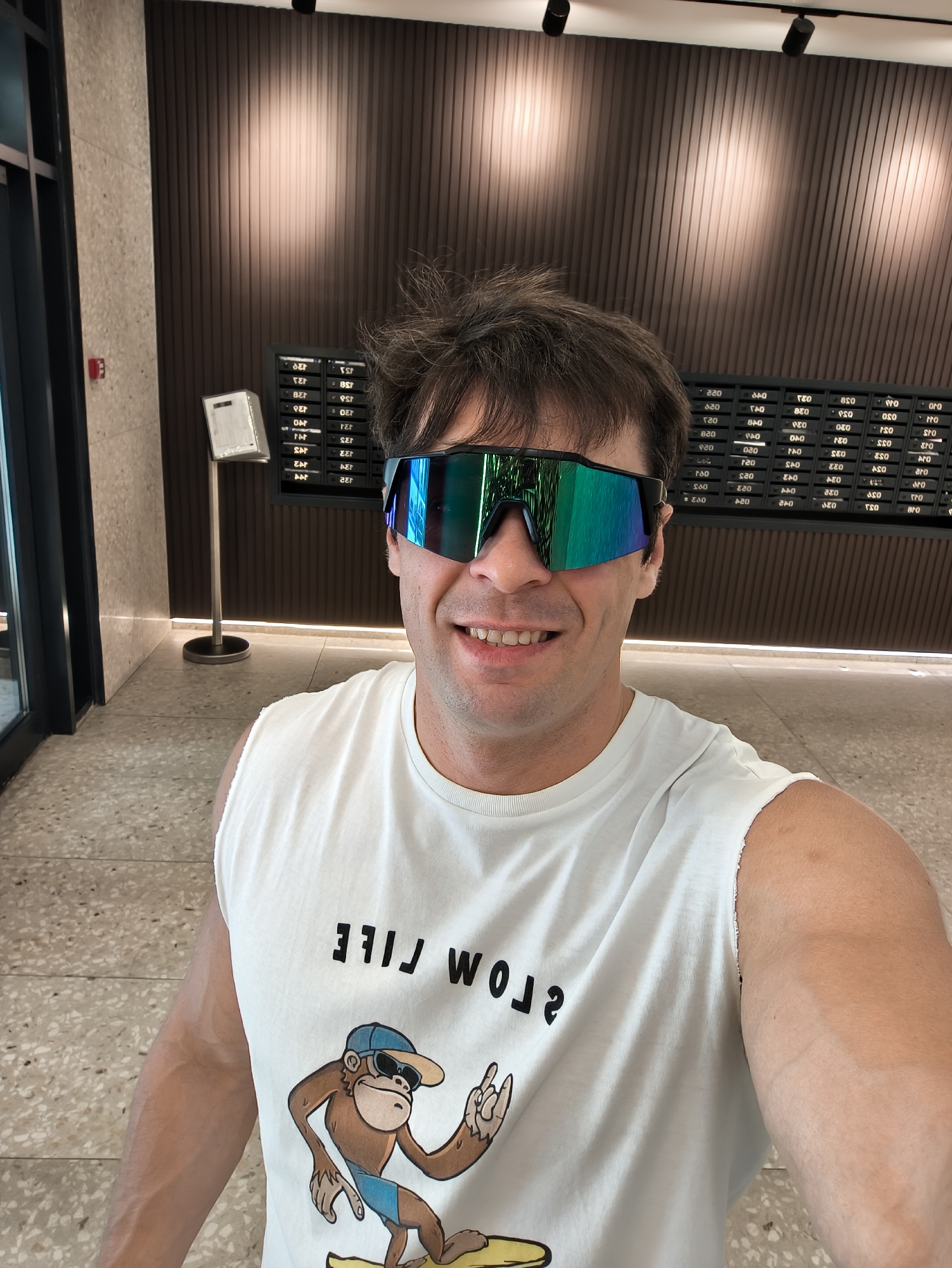 A man is taking a selfie indoors in what appears to be a residential lobby. He is wearing a white tank top with a graphic of a monkey on a surfboard and the words "SLOW LIFE" printed backward. He is also wearing mirrored sunglasses. Behind him, there is a wall with mailboxes and a directory. The floor is tiled. There is a door on the left and ceiling lights. The mailboxes have unit numbers on them, such as "123" and "156." The man's hair is messy. There is a small white message board on a stand to the left of the mailboxes. The overall lighting is bright.