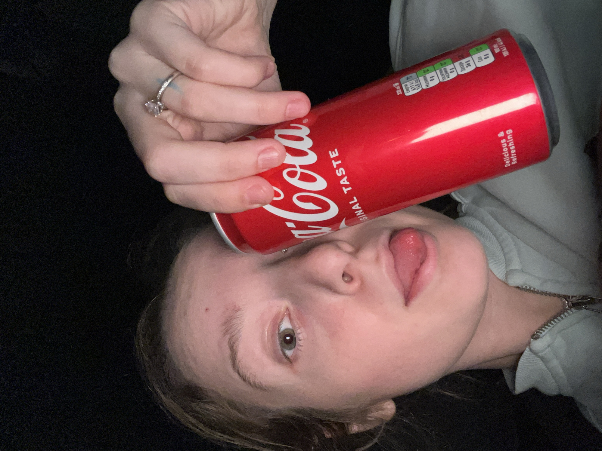 A woman lies on a dark surface, holding a red Coca-Cola can against her forehead with her hand, which also displays a diamond ring. Her tongue is slightly protruding. She is wearing a light-colored hooded sweatshirt. The photo has been taken from a tilted perspective. No information suggests the image was taken in Varna, Bulgaria.